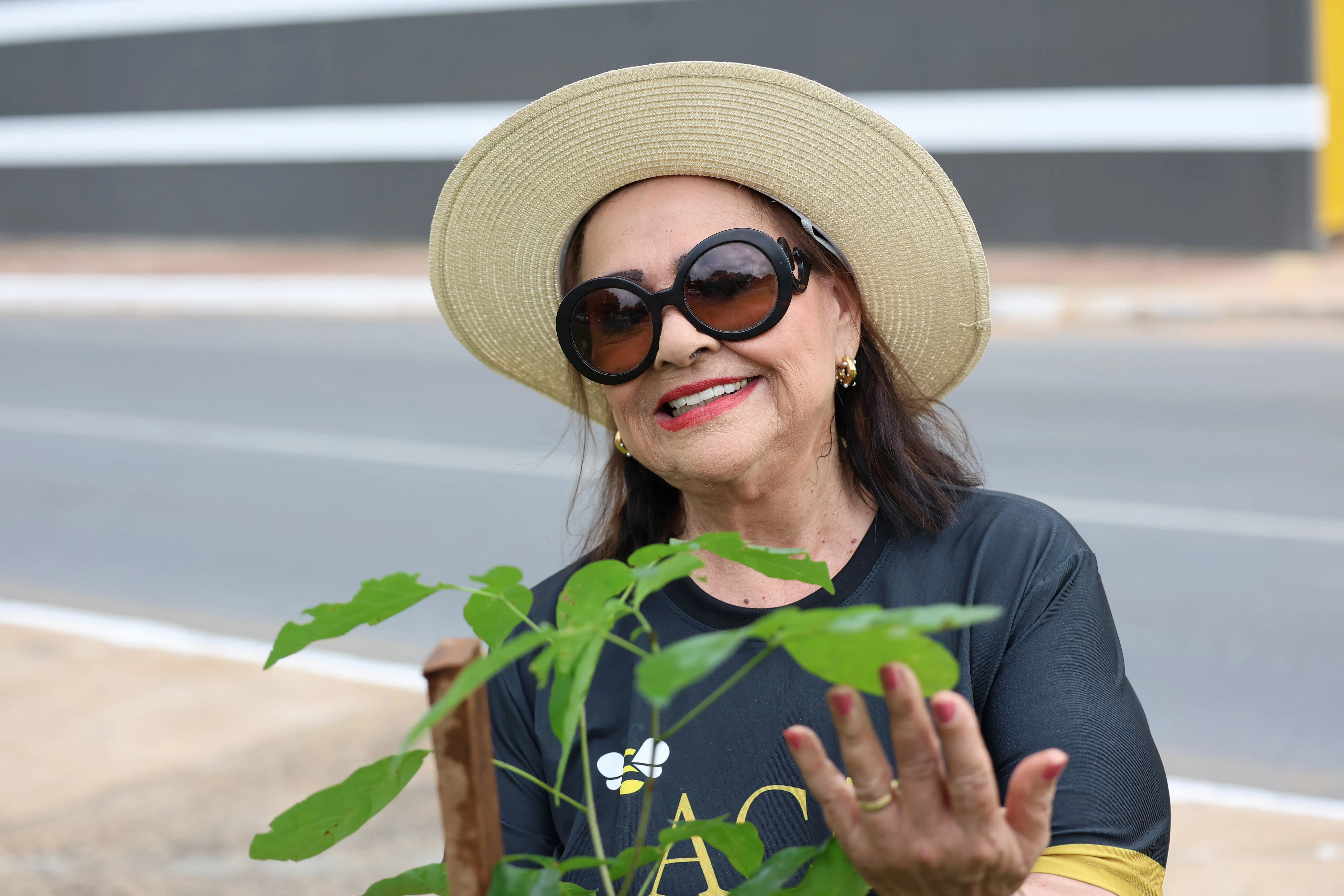 Mulher idosa sorri usando chapéu bege, óculos escuros arredondados e camiseta preta 
