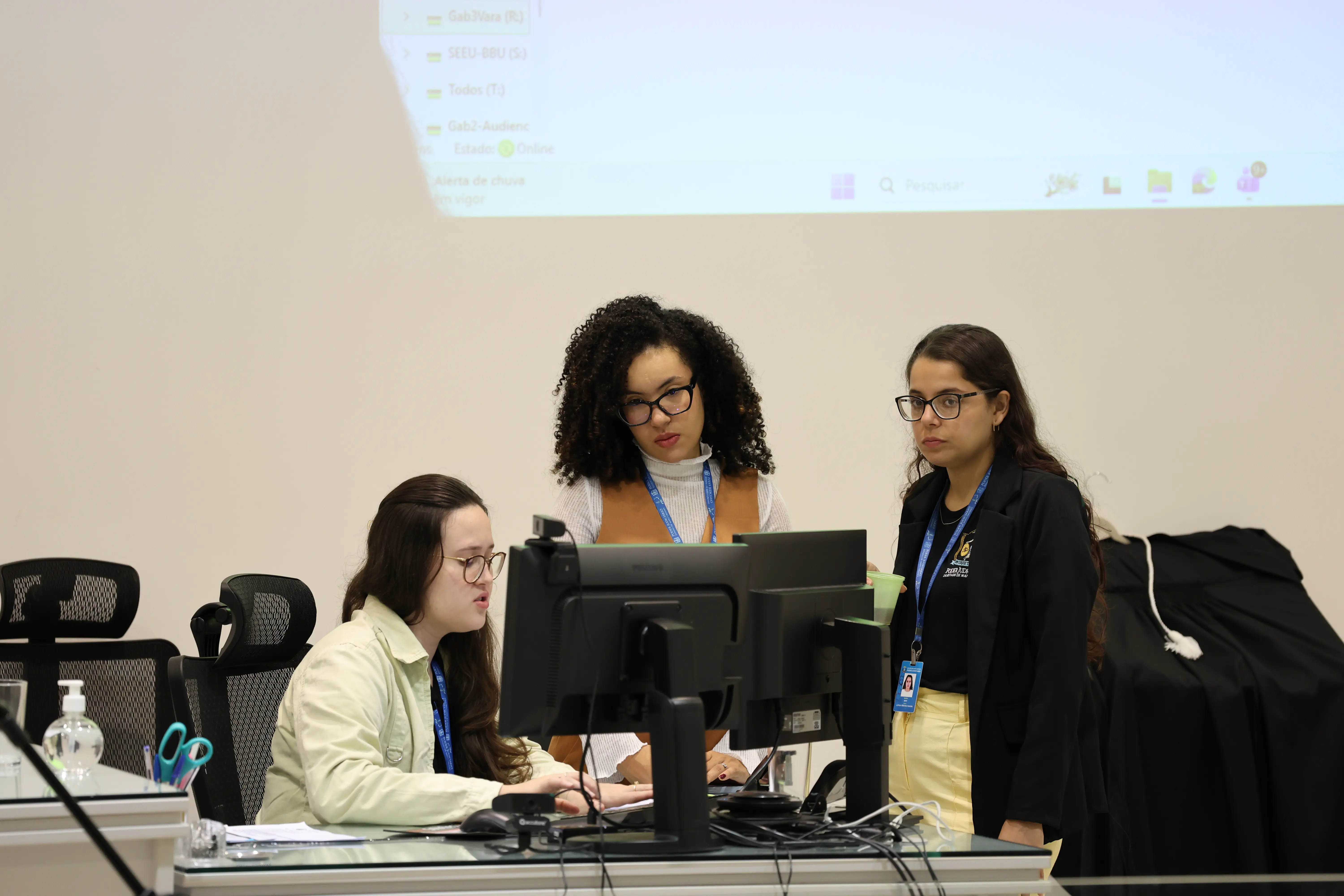 Três mulheres em estação de trabalho; duas sentadas e uma em pé. Observam telas de computador em sala iluminada.