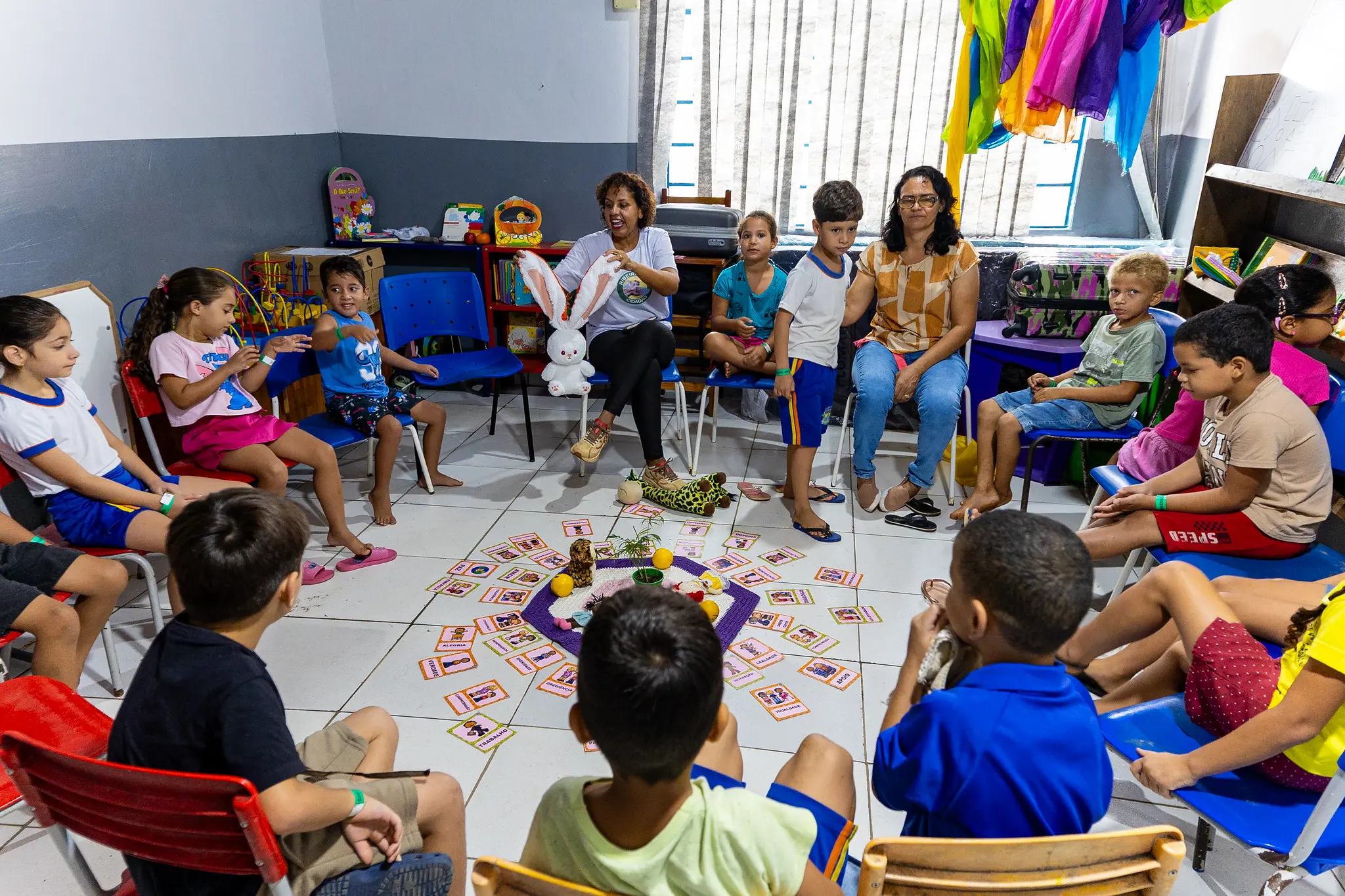 Sentadas em círculo, alunos da Escola Municipal Santo Antônio, no Distrito de Caramujo, município de Cáceres, participam das práticas restaurativas dentro de uma biblioteca escolar. Dispostos no centro do círculo, estão materiais incluindo cartões ilustrados e objetos lúdicos.