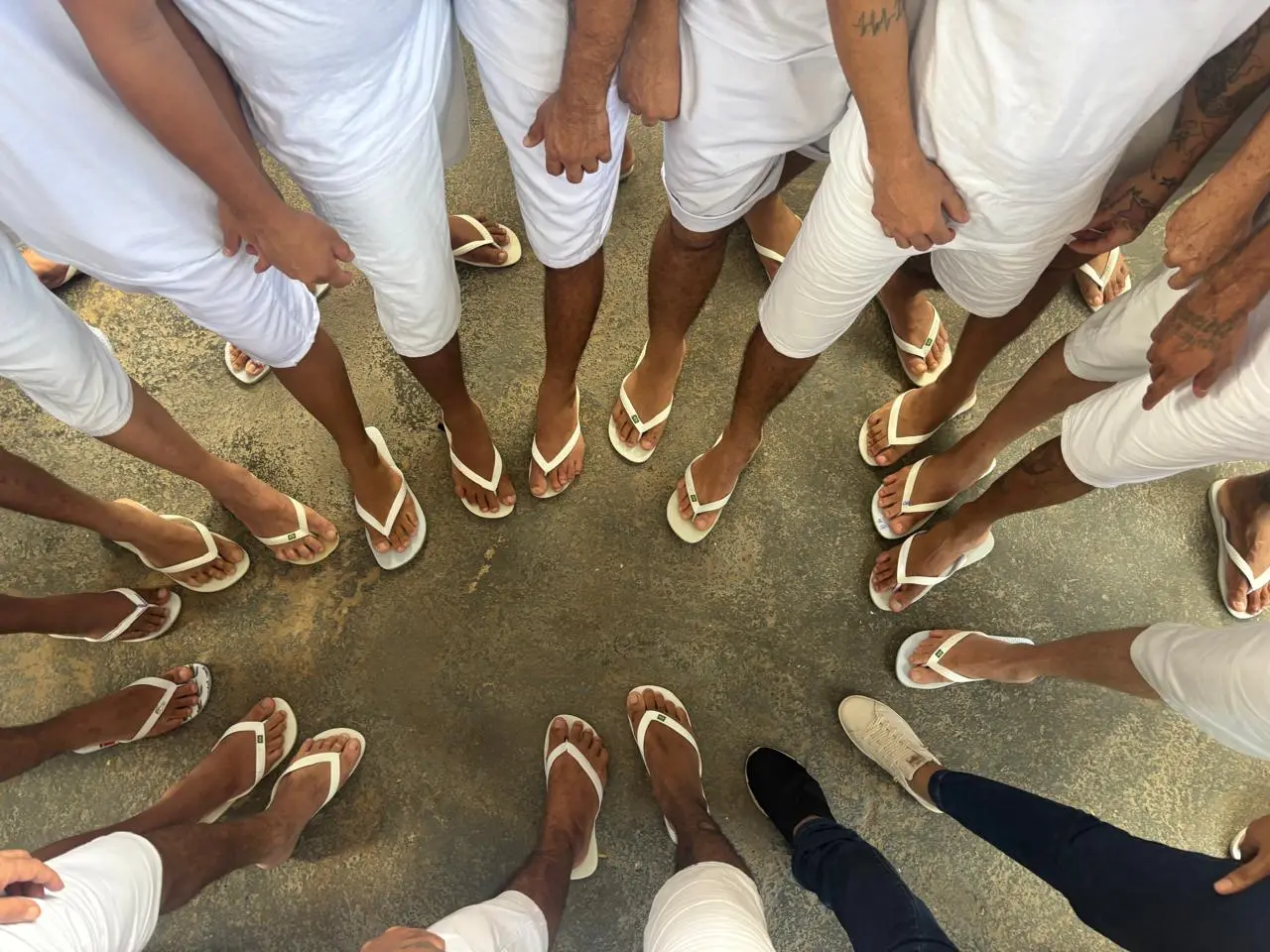 Imagem de cima para baixo, no qual aparece somente parte do copo dos participantes do Grupo Reflexivo para Homens, eles todos estão de pé, usam uniforme e chinelo branco, formando um círculo. As duas facilitadoras também colocaram seus pé, elas usam tênis, no círculo.