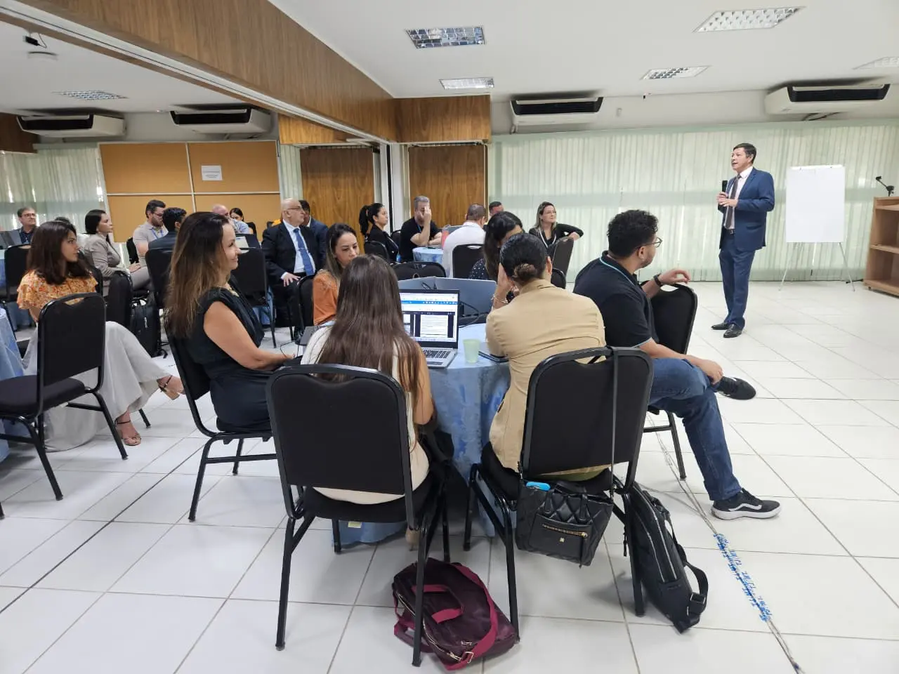 Foto colorida e horizontal de treinamento. Desembargador Lídio Modesto está em pé, à direita. Ele segura microfone e fala para plateia de aproximadamente 40 pessoas sentadas em cadeiras em um semicírculo.  Muitos dos participantes estão olhando para o palestrante, e há mesas e notebooks visíveis.