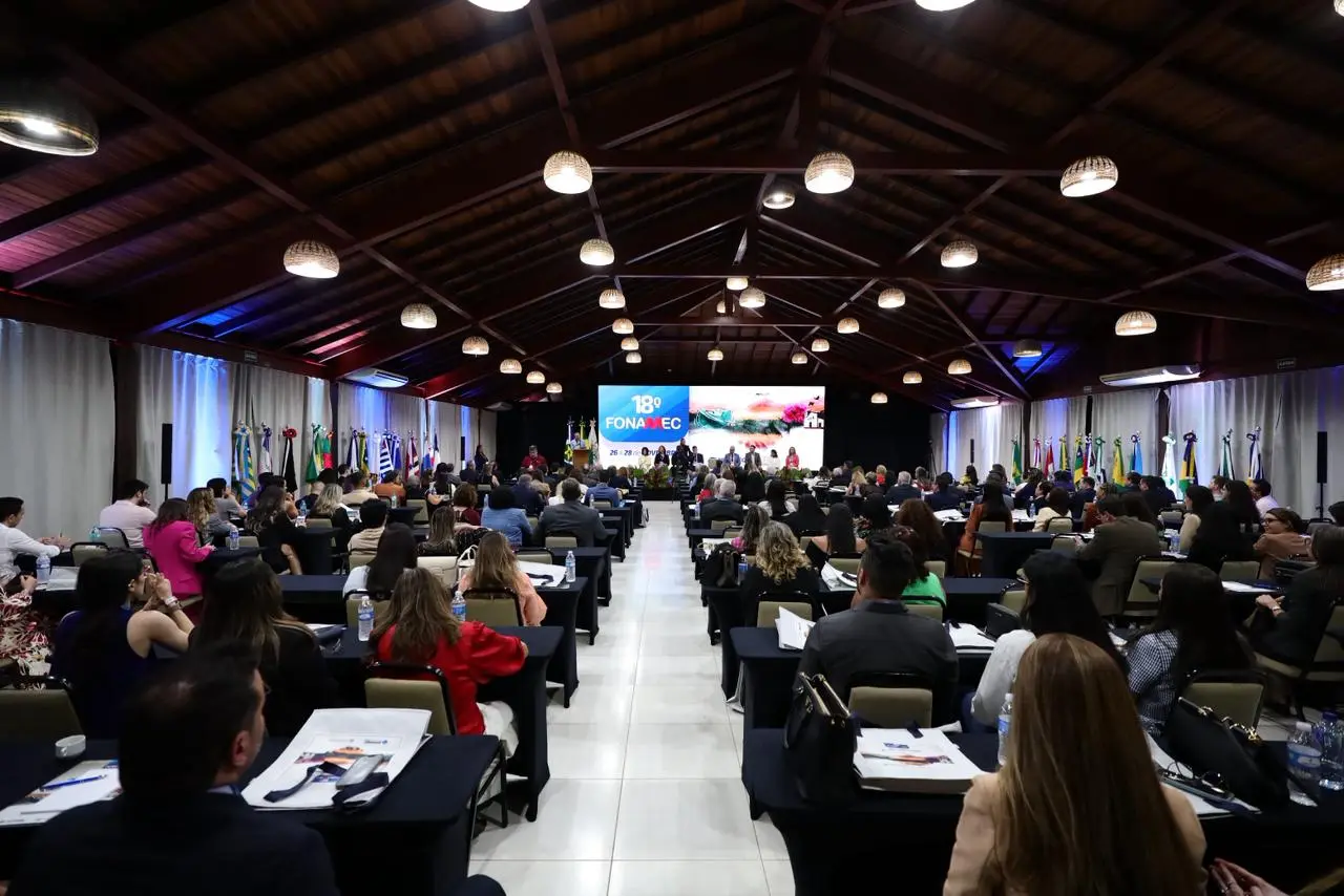 Foto em plano aberto que mostra um auditório lotado de pessoas sentadas, participando do Fonamec. No palco, há várias autoridades em pé.
