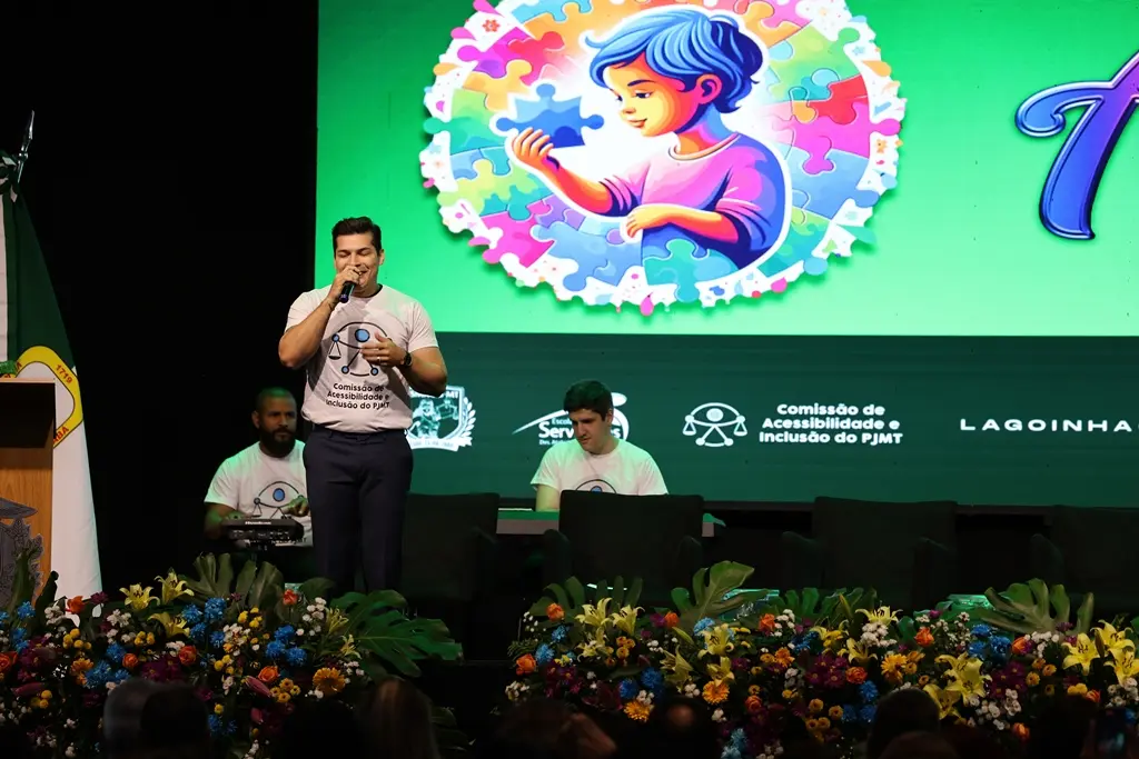 Foto do cantor Tiago Roseno cantando no palco do evento TJMT Inclusivo, Atrás dele, há dois músicos o acompanhando: um percussionista e um pianista. Os três usam a camiseta branca com o símbolo universal da acessibilidade.