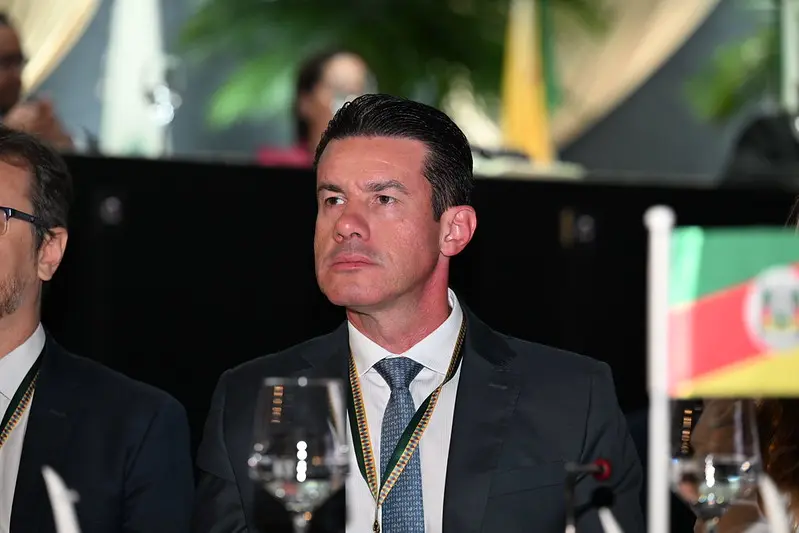 Desembargador Rodrigo Curvo sentado à mesa de debates do Colégio de Ouvidores Judiciários. Ele é um homem branco, magro, de olhos e cabelos escuros, usando camisa branca, paletó preto, gravata azul e crachá do evento.