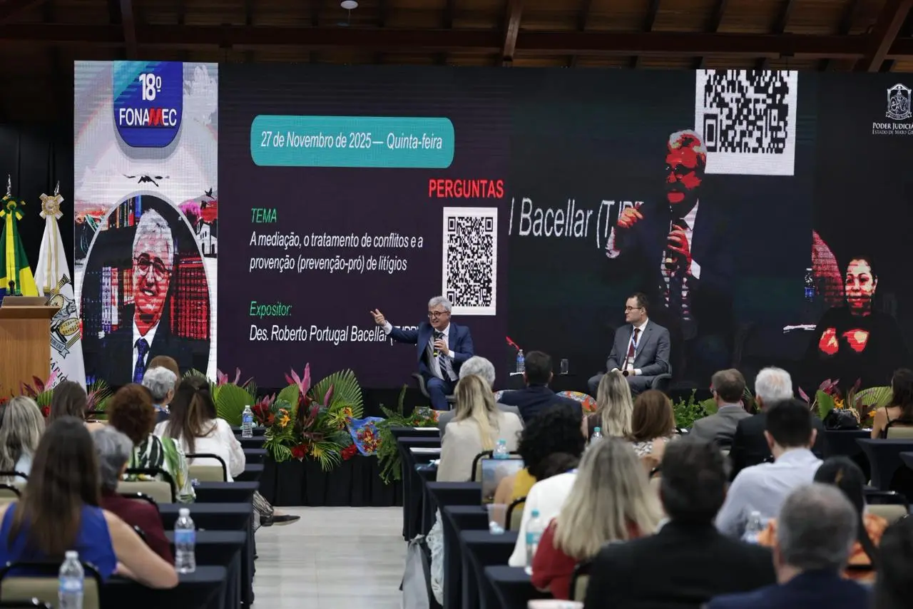 Foto em plano aberto que mostra o auditório lotado de pessoas assistindo a palestra de Roberto Bacellar, que está falando ao microfone, no palco. ao lado dele, está o mediador do painel, juiz Eduardo Peruffo.