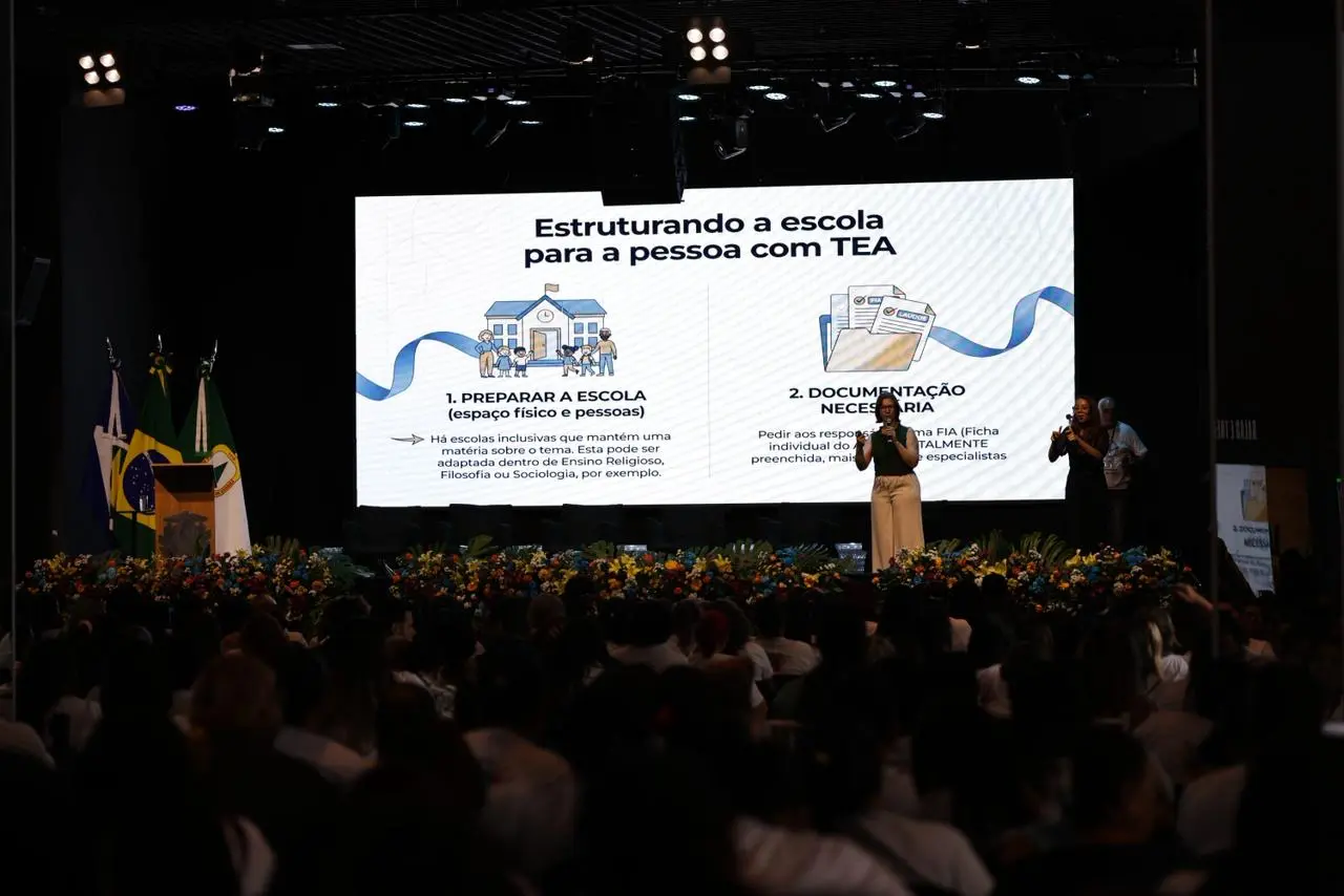 A imagem mostra o auditório lotado enquanto a palestrante apresenta conteúdo no telão sobre estrutura escolar para pessoas com TEA. Bandeiras estão à esquerda do palco e flores coloridas decoram a frente.