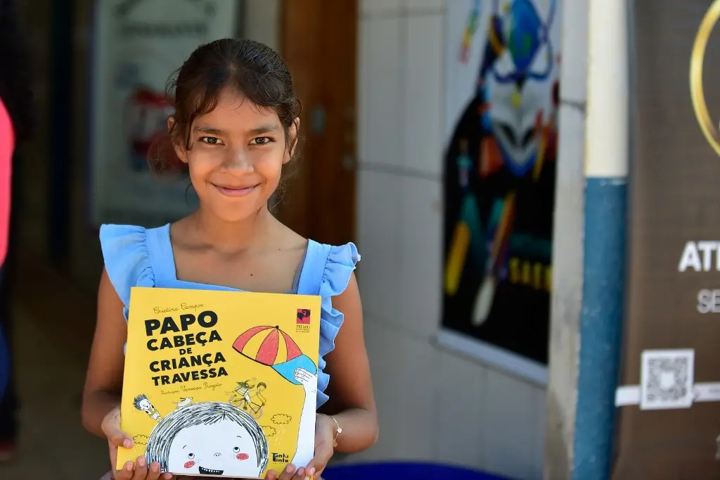 Menina sorri para a foto, mostrando um livro intitulado "Papo Cabeça de Criança Travessa". Ela está no pátio de uma escola. ela é negra, de olhos castanhos, cabelos lisos, castanhos e presos, usando vestido azul claro.
