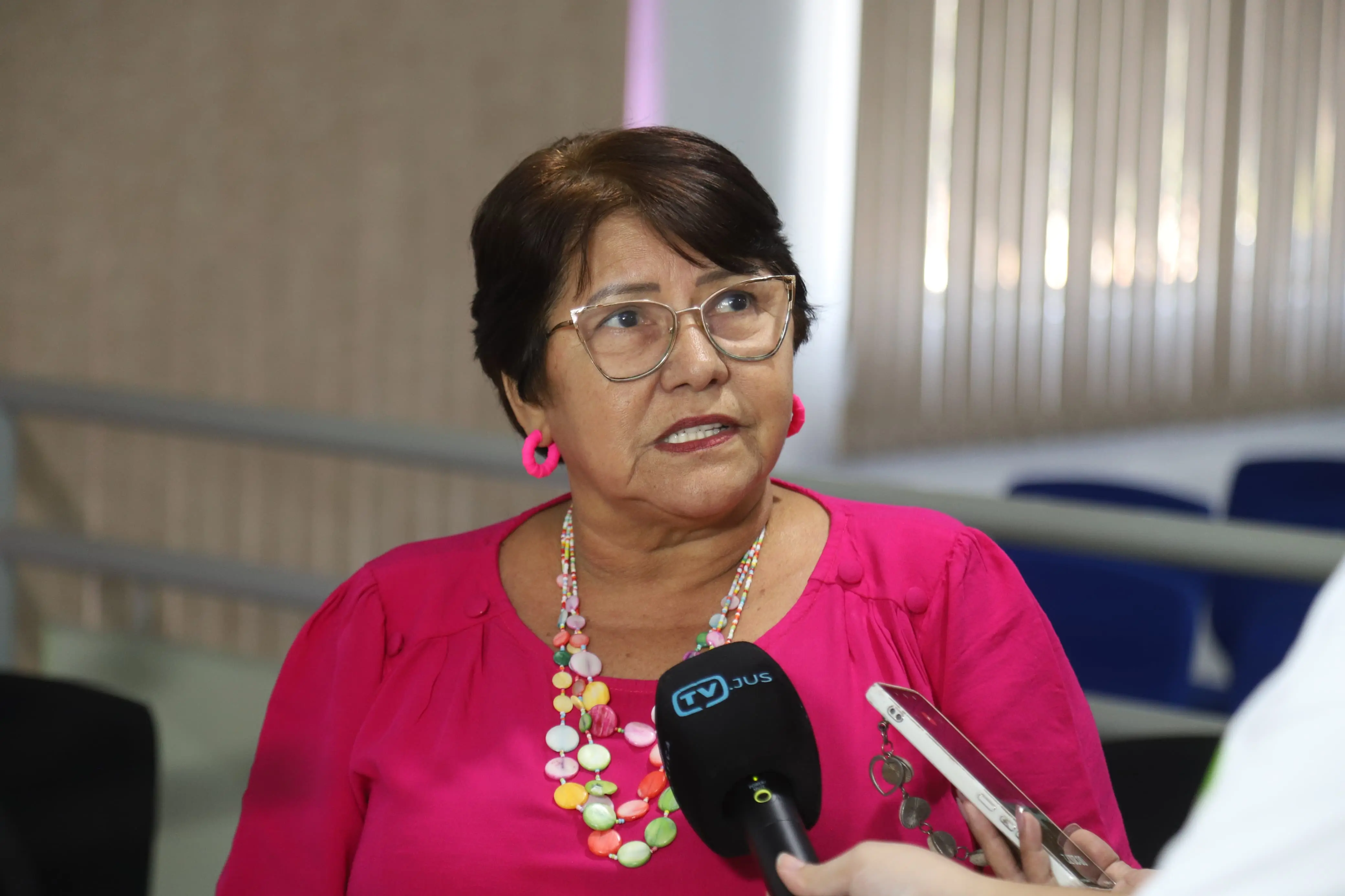 Mulher de óculos, com blusa rosa e colar colorido, dá entrevista segurando microfone da TV Justiça. Ela usa brincos rosa e tem expressão séria. Ao fundo, cadeiras azuis e persianas claras, em ambiente de evento institucional.