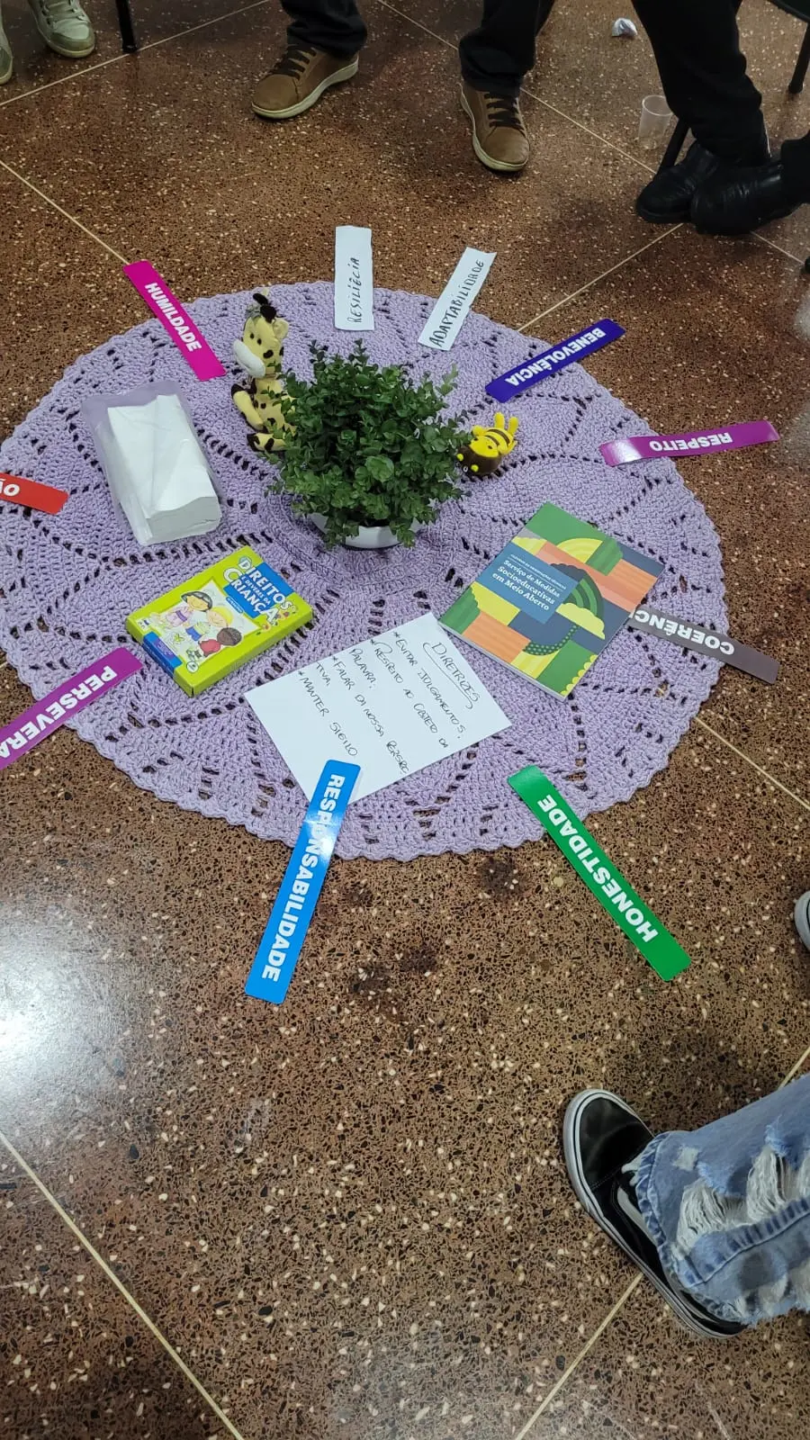 Uma vista aérea de um círculo de crochê lilás no chão, decorado com palavras coloridas como "Respeito" e "Humildade". Há também um vaso de plantas, lenços e livros. Pés de pessoas ao redor.