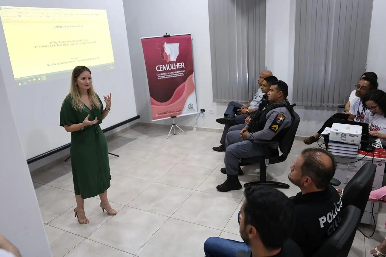 apresentação em ambiente interno, com a mesma palestrante de vestido verde se dirigindo a grupo misto de policiais militares e civis sentados em semicírculo, equipamento de projeção ao centro.