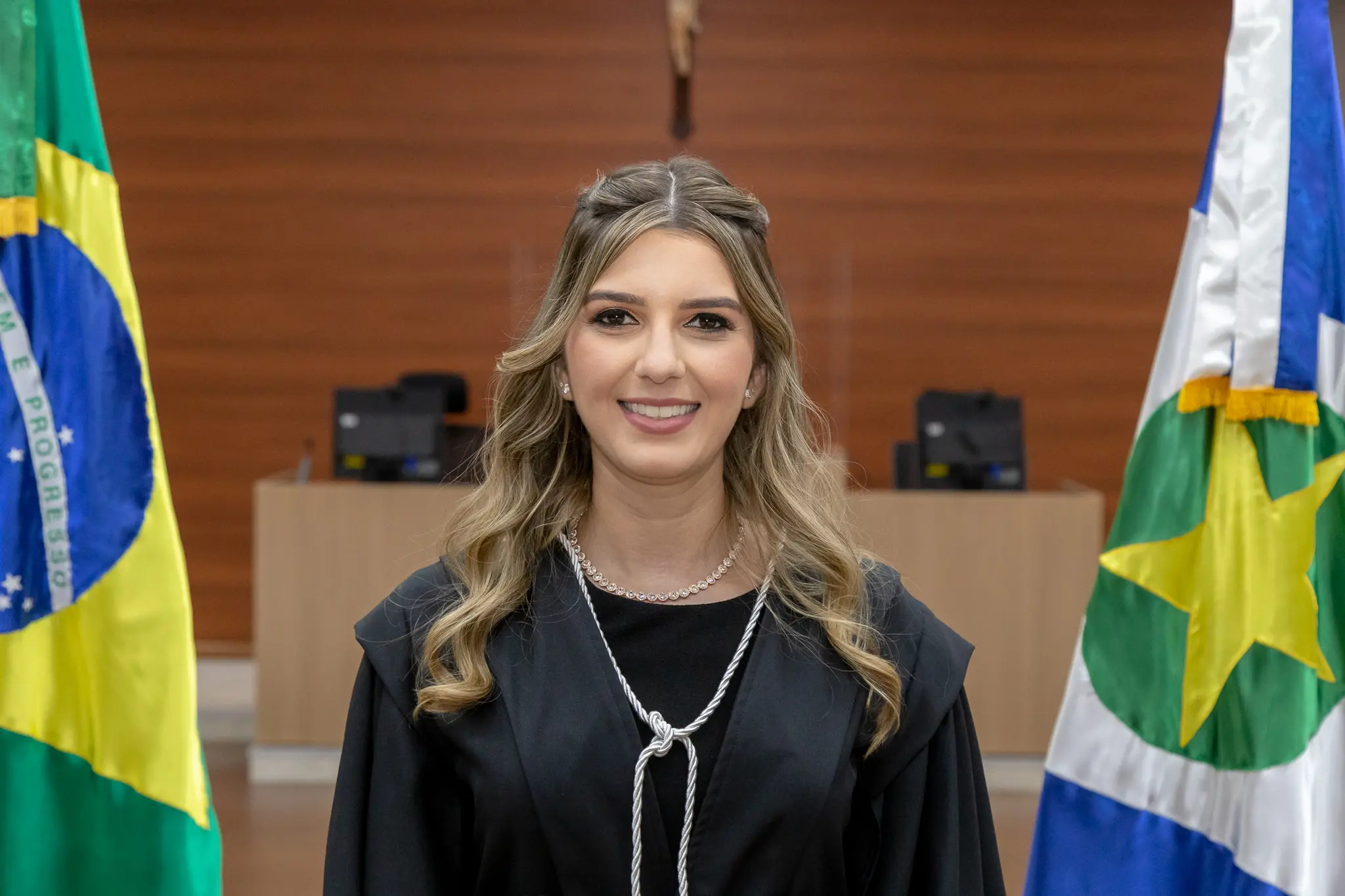 Foto de Amanda Dias uma jovem mulher de pele clara, com cabelos loiros ondulados e compridos, parcialmente presos com tranças nas laterais. Ela sorri levemente e olha diretamente para a câmera. Veste uma beca preta, uma camisa branca por baixo com uma espécie de jabô.