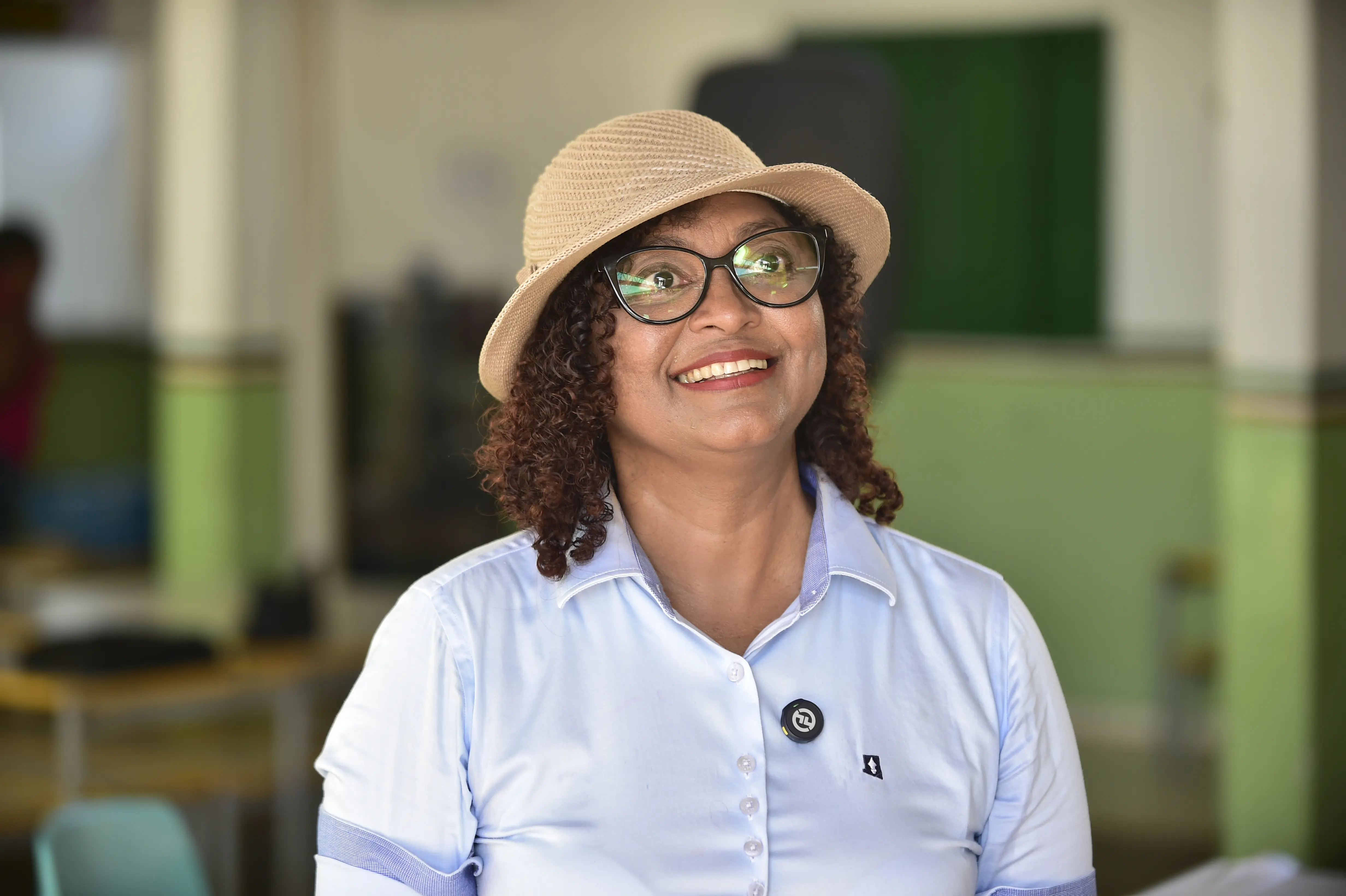 Uma mulher sorridente com cabelo cacheado escuro, usando óculos e um chapéu de palha claro. Ela veste uma camisa azul clara e um broche preto no peito. O fundo está desfocado.