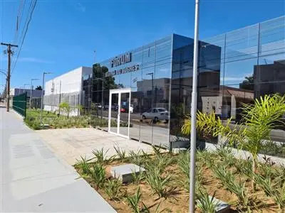 O exterior de um moderno edifício, provavelmente um fórum, com fachada de vidro espelhado. Em destaque na fachada, o nome "FÓRUM BARRA DO BUGRES". Em frente, um jardim com plantas baixas e palmeiras jovens. Um dia ensolarado, com reflexos no vidro e uma calçada à esquerda.