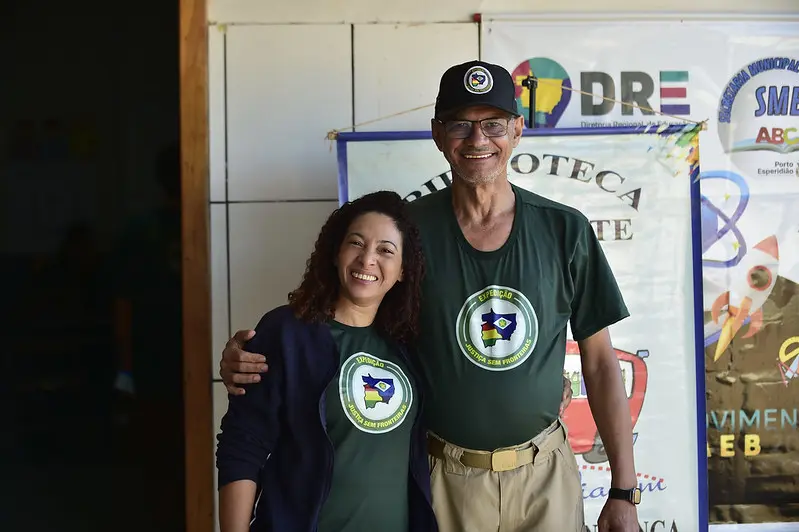 Uma mulher e um senhor se abraçam de lado e sorriem para a foto, no pátio de uma escola. Ambos usam a camiseta verde musgo do projeto Justiça Sem Fronteiras.