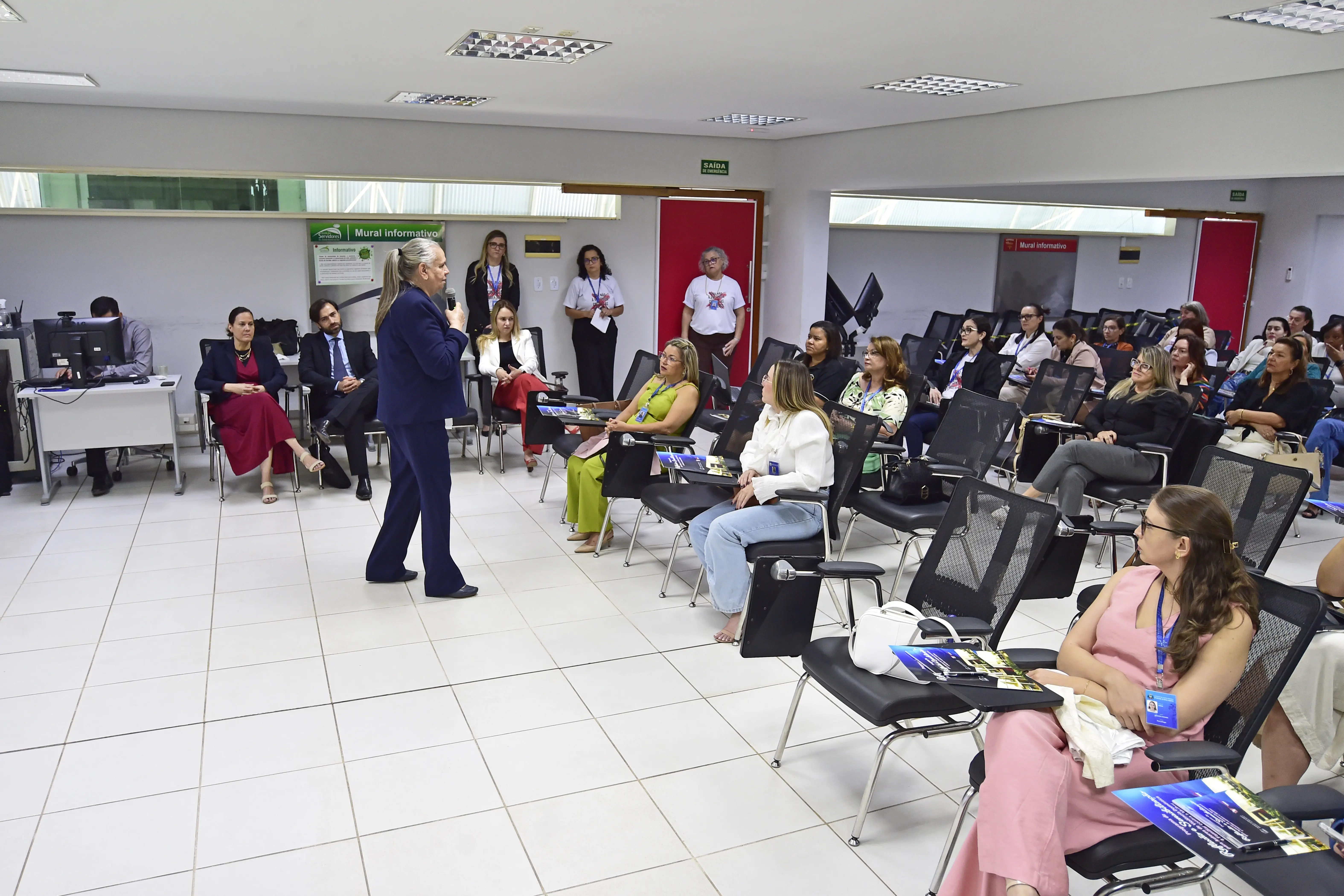 Foto em plano aberto que mostra a sala de aula cheia de pessoas sentadas e a desembargadora Maria Erotides Kneio em pé, falando ao microfone.