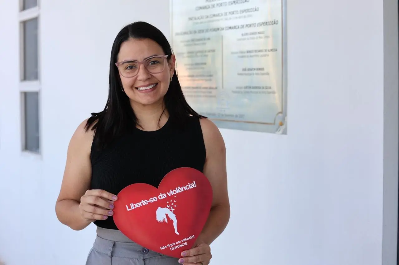 Foto posada de mulher de blusa amarela sorrindo e segurando a mesma placa vermelha em formato de coração com mensagem contra violência, em corredor interno.