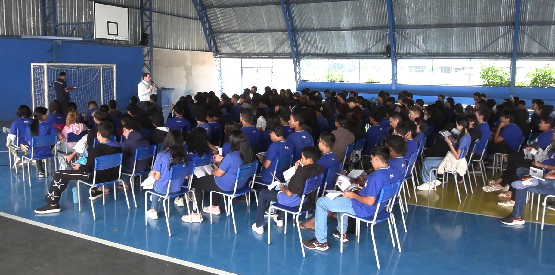 Foto em plano aberto que mostra uma quadra coberta da escola repleta de alunos uniformizados e sentados. Eles assistem à palestra do servidor do TJMT, que está à frente, em pé, falando ao microfone. 