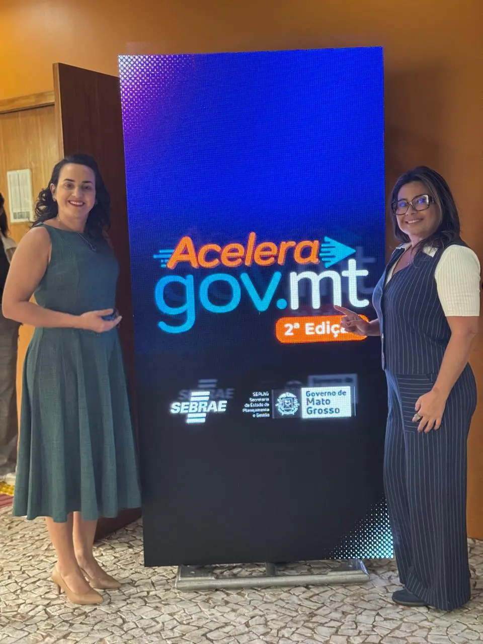Duas mulheres sorridentes flanqueiam uma tela vertical que exibe "Acelera Gov.mt 2ª Edição" em destaque, com logos do Sebrae e Governo de MT. Uma veste um vestido verde, a outra um macacão listrado.
