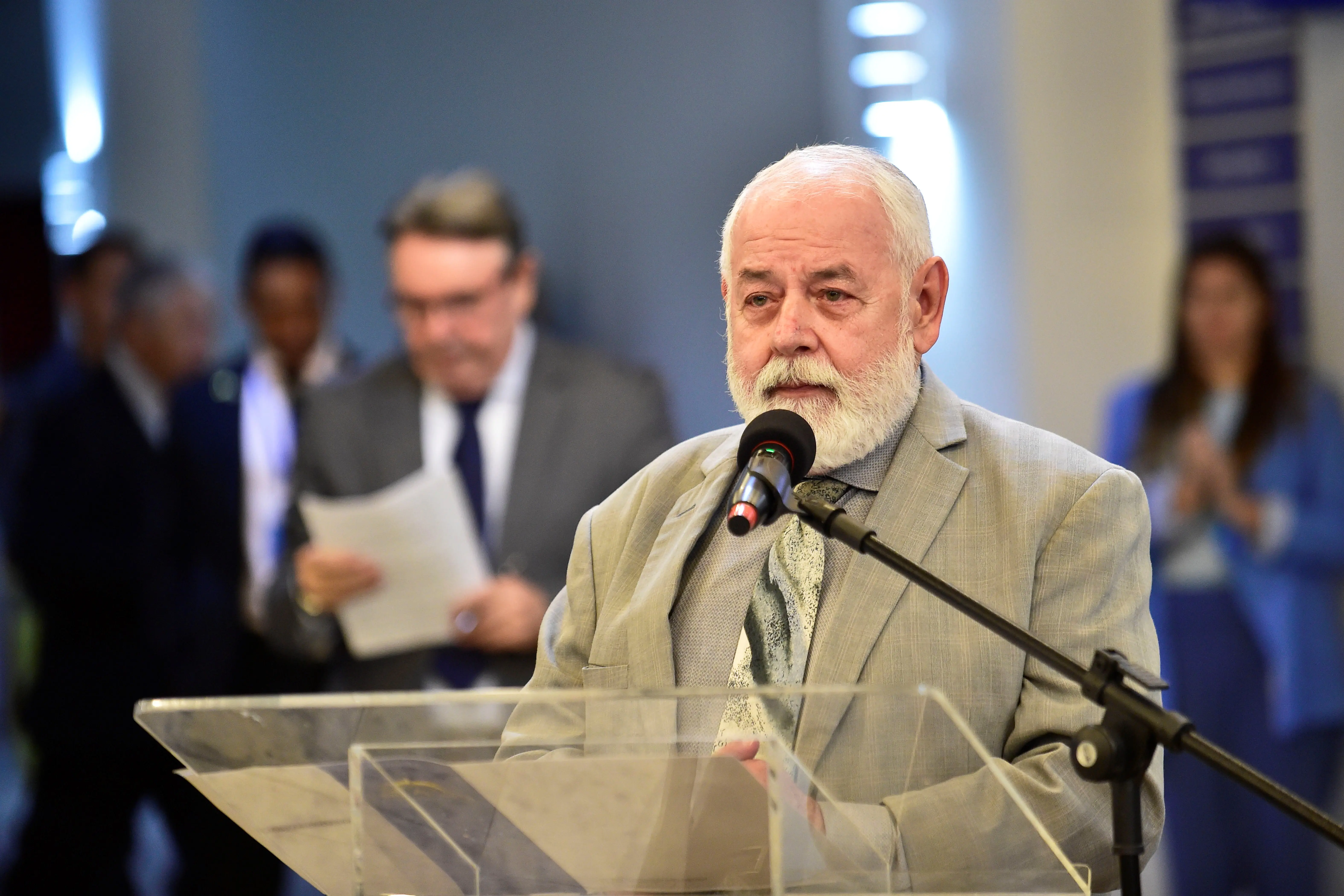 Na foto, um homem branco , com barba branca e cabelo grisalho está falando em um pódio de acrílico com um microfone. Ele usa um paletó cinza claro e uma gravata estampada.