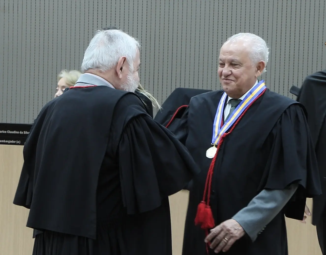 Foto que mostra o presidente do TJMT, desembargador José Zuquim, de costas, cumprimentando o desembargador Luiz Ferreira da Silva, que usa o colar do mérito judiciário. Ambos sorriem um para o outro e dão um aperto de mãos.
