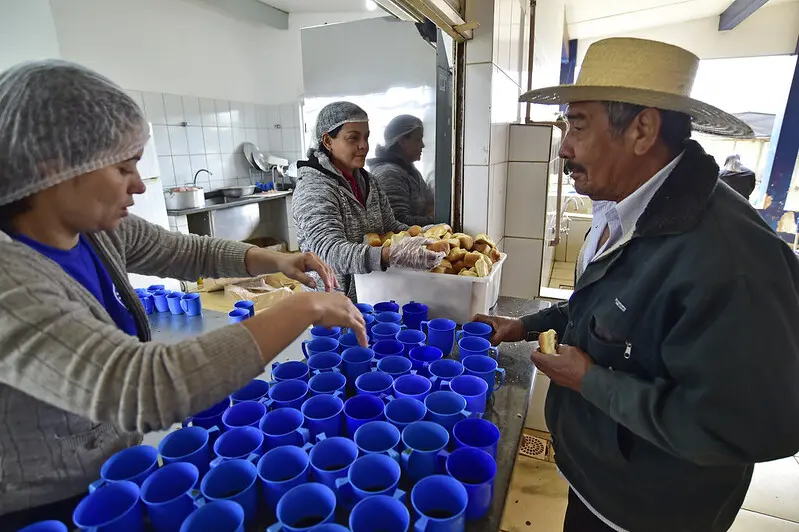 Foto que mostra uma merendeira servindo chá a um senhor e outra servindo pães, no balcão da cozinha da escola onde ocorreu o mutirão da Justiça Sem Fronteiras.