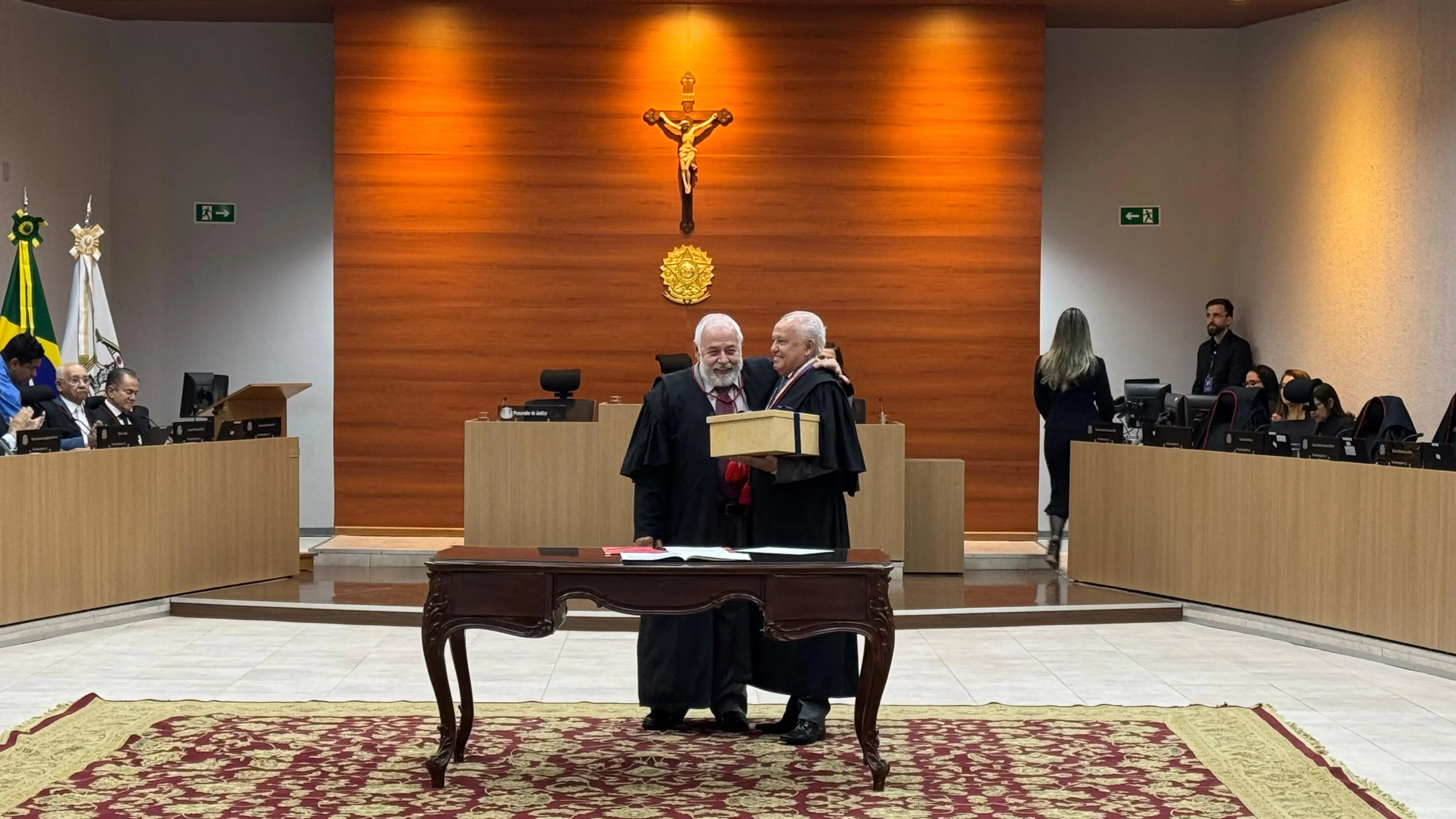 Foto em plano aberto que mostra o presidente do TJMT, desembargador José Zuquim Nogueira, e o desembargador Luiz Ferreira da Silva, ao centro do Plenário 1 do Tribunal, em pé e se abraçando. Ferreira segura uma caixa onde contém sua toga e demais objetos de trabalho.