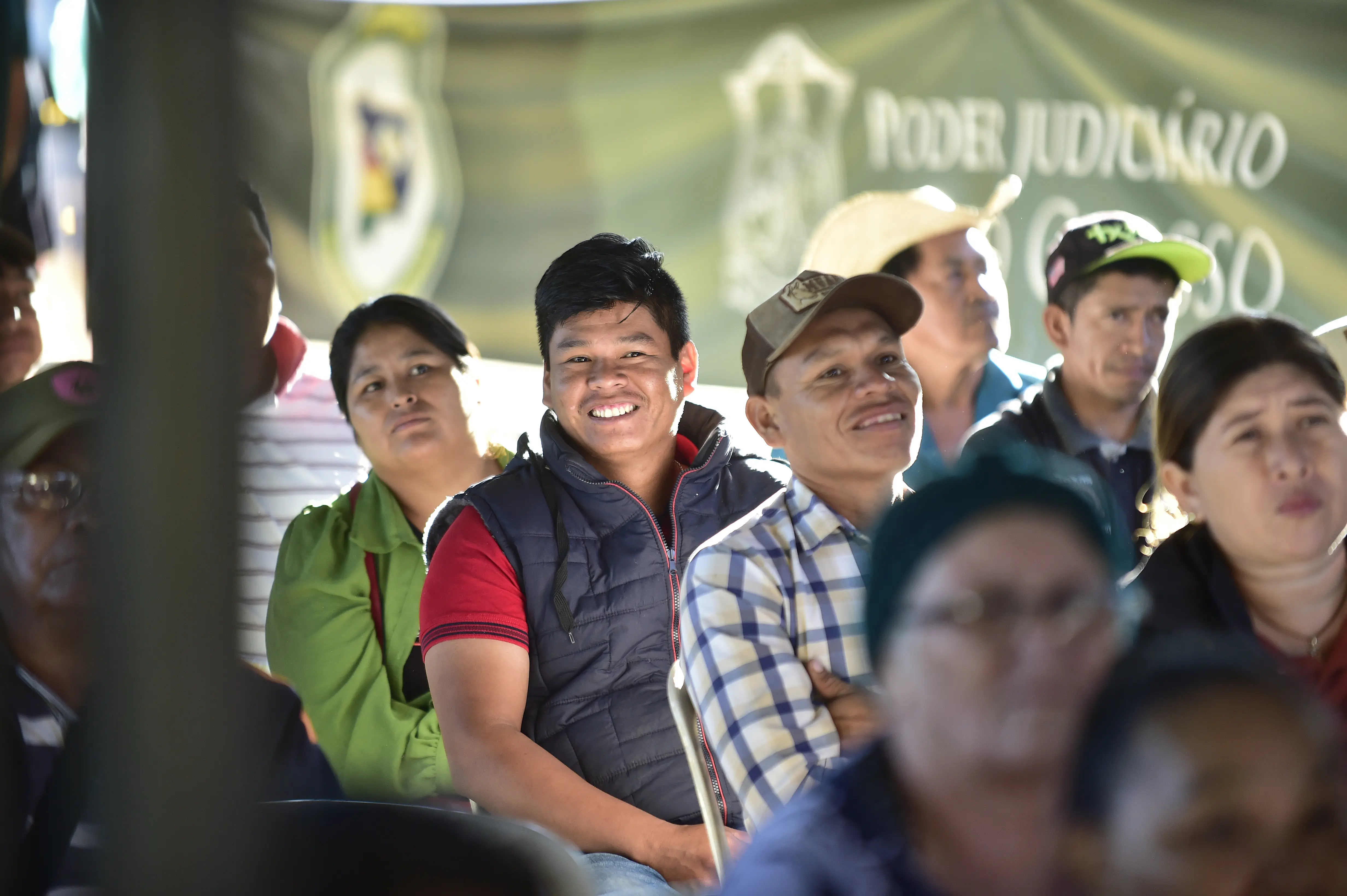 Grupo de pessoas assiste atentamente a uma atividade da Expedi��o Justi�a Sem Fronteiras. Em destaque, homem sorri em meio ao p�blico, que est� reunido sob uma tenda com banner do Poder Judici�rio ao fundo.