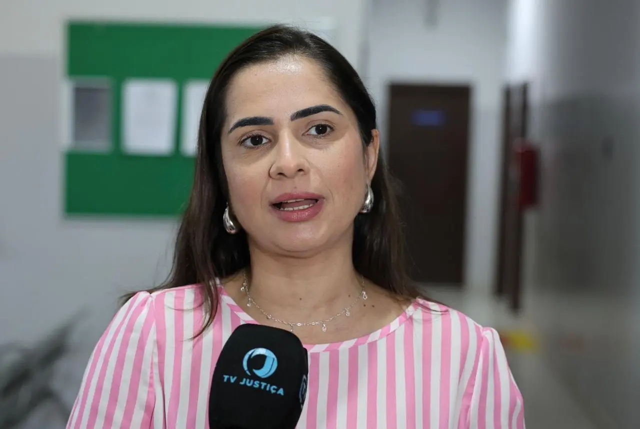 Entrevista para TV Justiça com mulher de blusa rosa listrada falando ao microfone em ambiente interno com quadros informativos ao fundo.