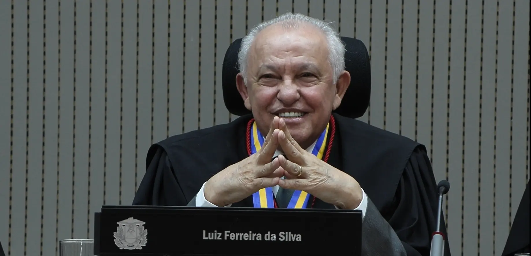 Desembargador Luiz Ferreira da Silva sentado em seu assento no Tribunal Pleno, sorrindo, com as mãos juntas. Ele é um senhor de pele e cabelos brancos, usando toga. 