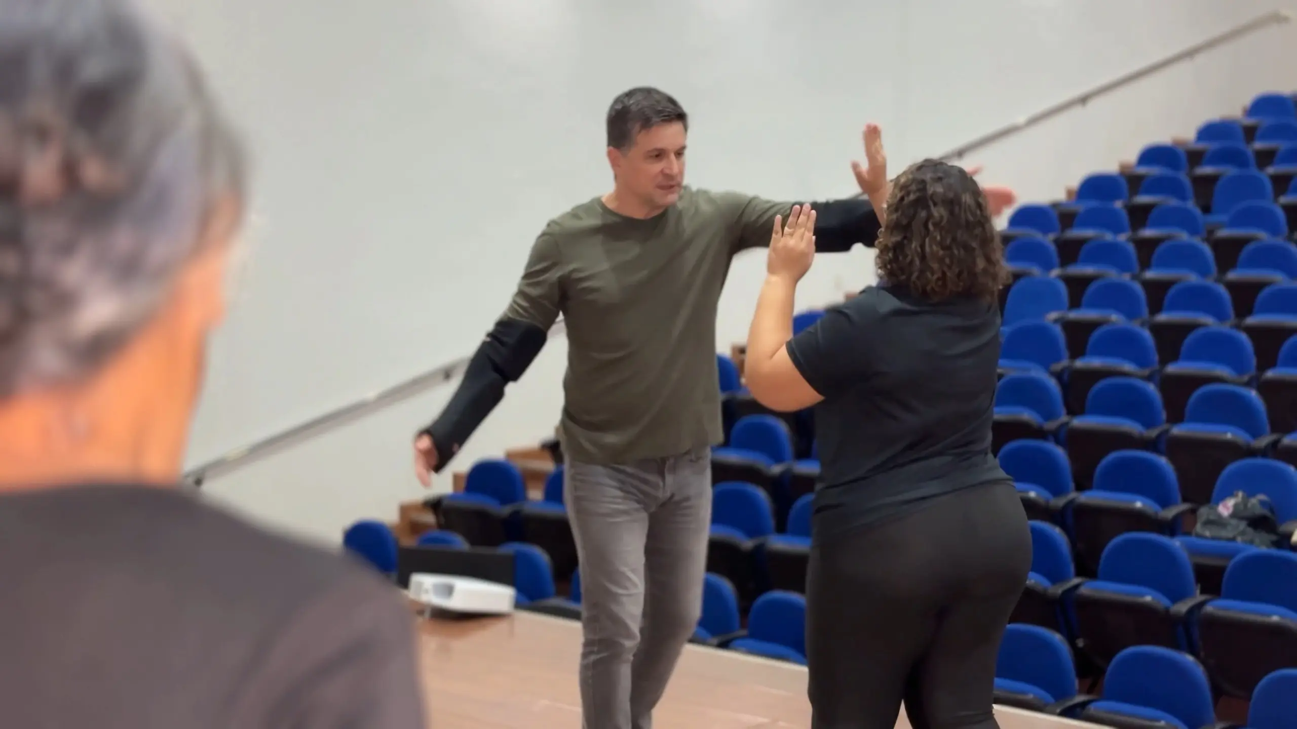 Nesta imagem, um homem e uma mulher interagem em primeiro plano.O homem, com cabelos curtos e grisalhos e camiseta verde-oliva, está de frente para a mulher, com o braço esquerdo estendido à altura do ombro dela