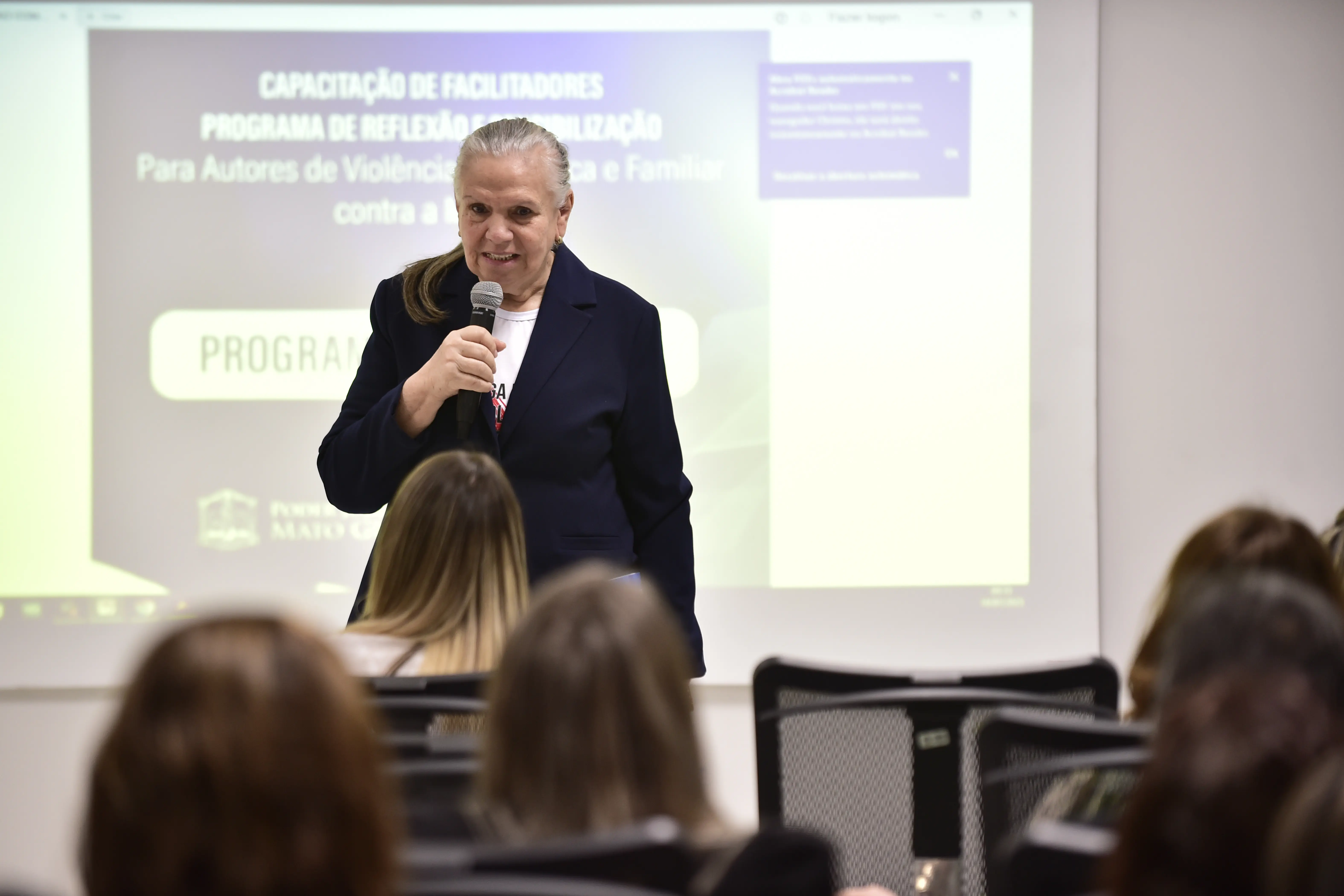 Foto em plano aberto que mostra a desembargadora Maria Erotides falando ao microfone, de frente para as pessoas que estão sentadas, de costas na foto. Atrás da desembargadora, aparece um telão com as informações do curso. A magistrada é uma senhora branca, de cabelos longos, lisos e grisalhos.
