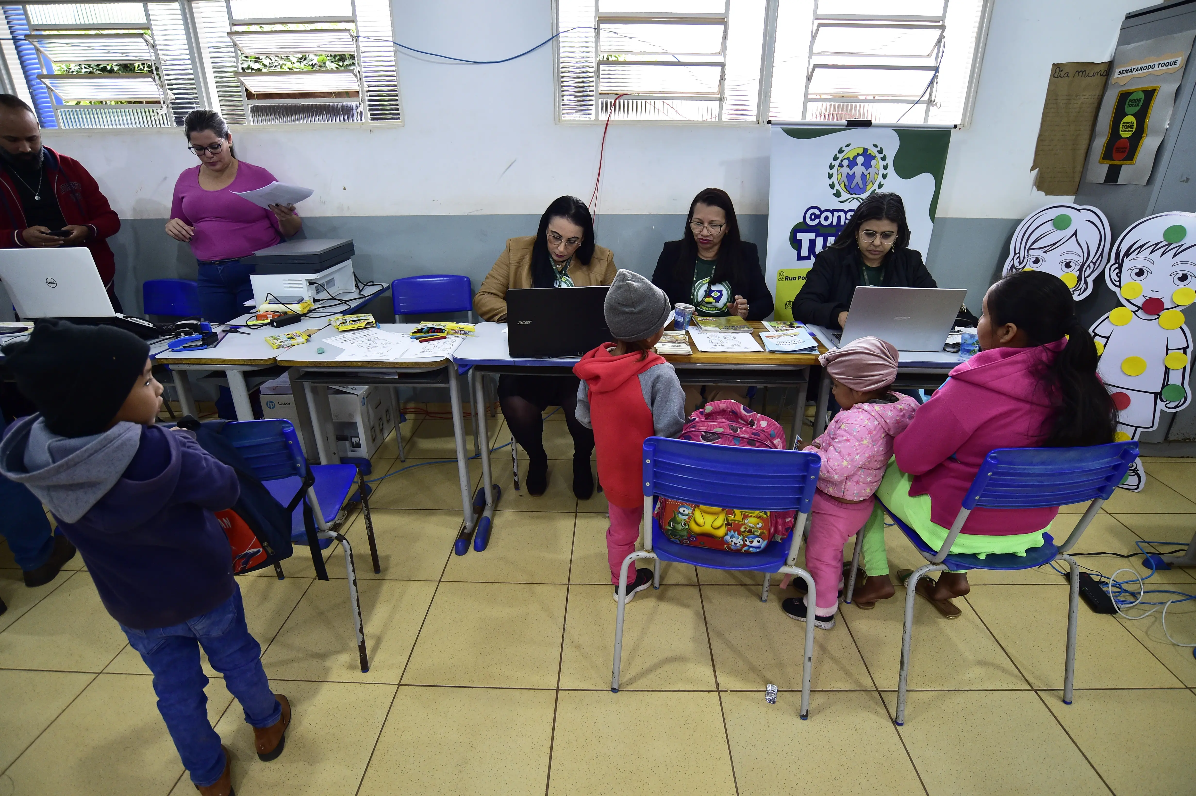 Três mulheres atendem uma mãe com duas meninas pequenas vestidas para o frio. Um menino observa a cena. Ao fundo, banner “Conselho Tutelar” e desenhos infantis decoram o local.