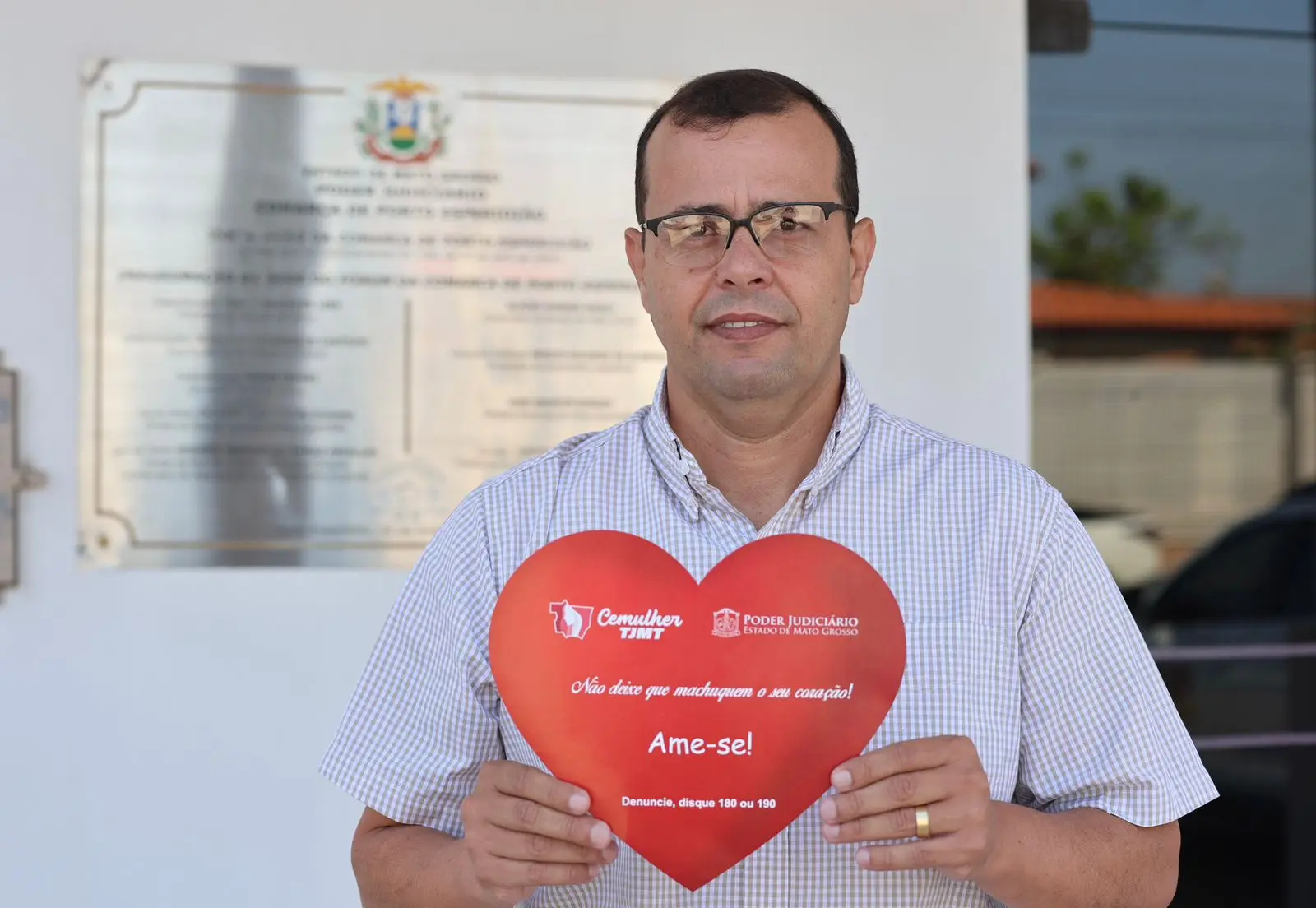 Homem de camisa xadrez azul e óculos posa em área externa segurando placa vermelha em forma de coração com mensagem "Não deixe que machuquem o seu coração! Ame-se!" e orientação para denúncia, com documento oficial ao fundo