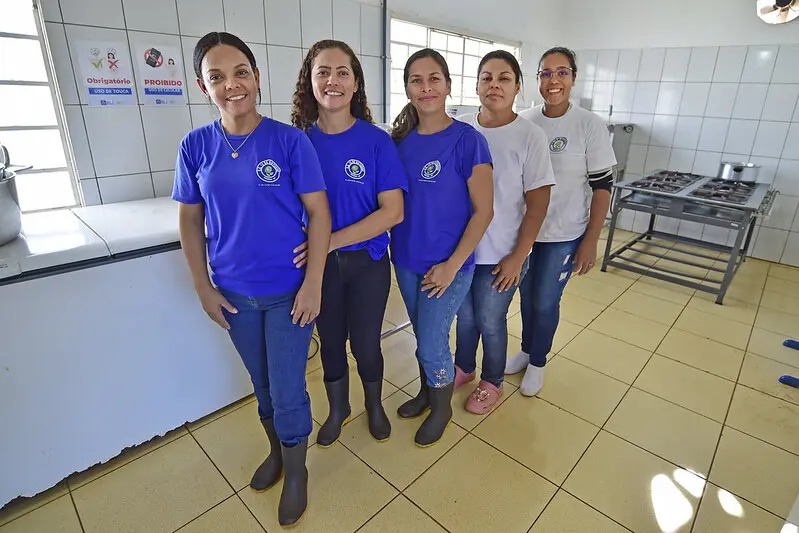 Foto que mostra cinco mulheres que trabalham na Escola Estadual 12 de Outubro posando para a foto e sorrindo. Elas estão em pé, na cozinha da escola.
