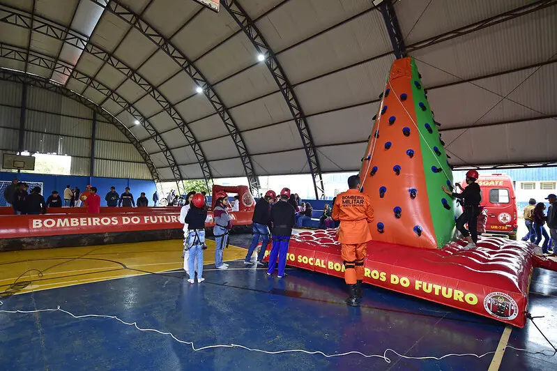 Foto que mostra a quadra poliesportiva da escola repleto de crianças e adolescentes brincando na torre de escalada e no campo inflável do Corpo de Bombeiros Militar.