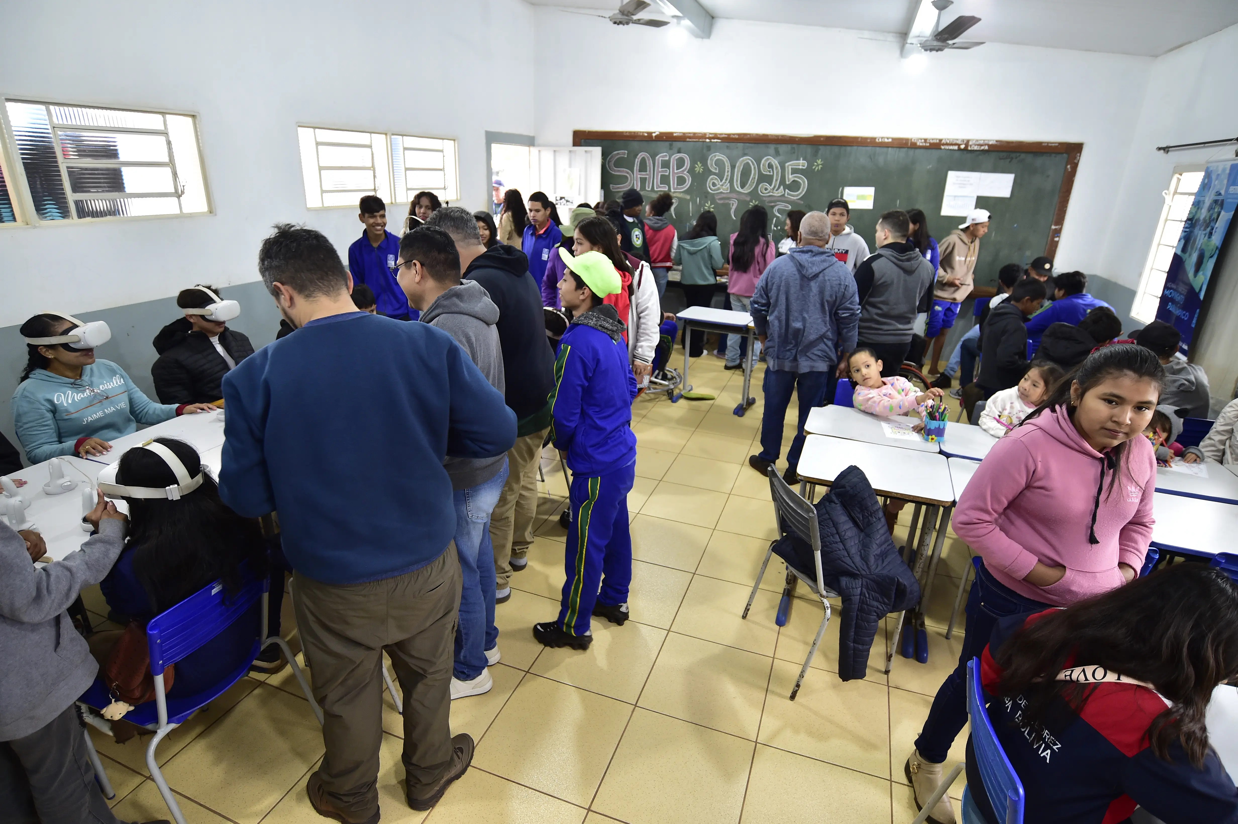 Sala de aula movimentada com adultos, jovens e crianças. Alguns usam óculos de realidade virtual. No fundo, lousa escrita "SAEB 2025". Crianças desenham e interagem em mesas escolares.