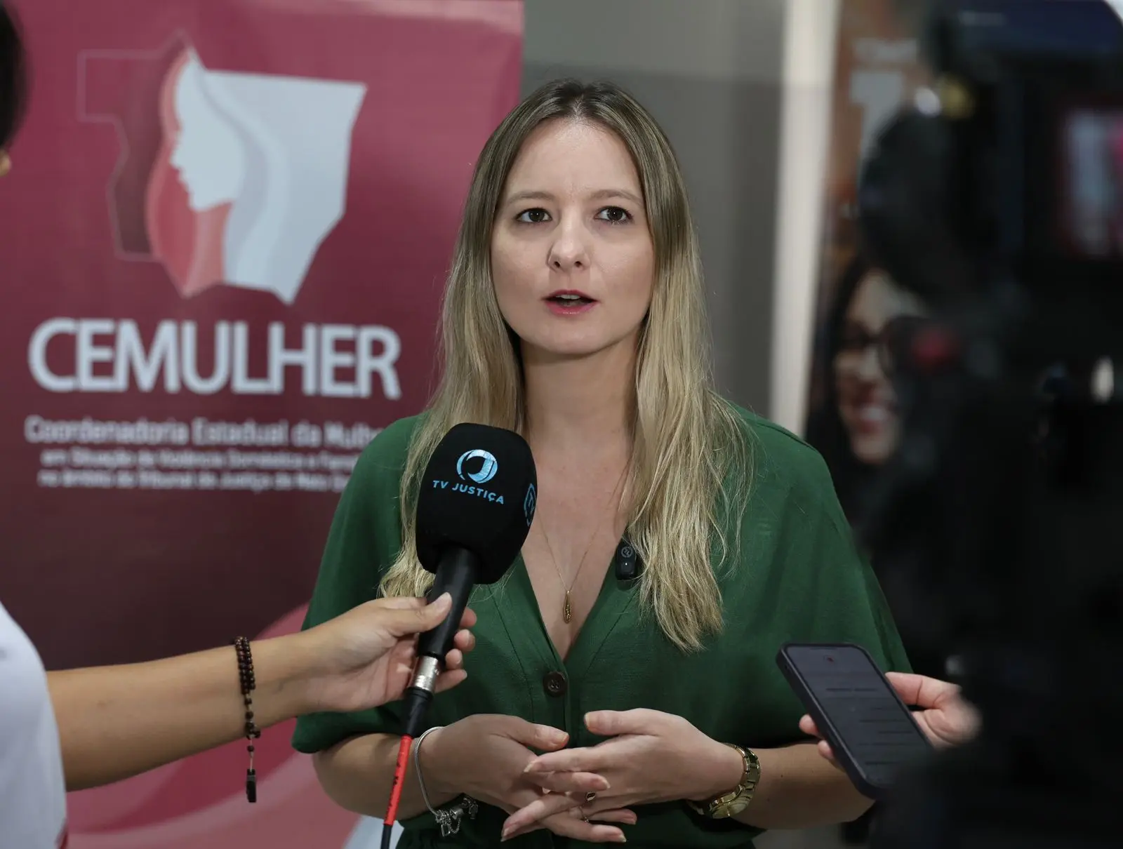 Entrevista coletiva com mulher de vestido verde respondendo a jornalistas da TV Justiça e outros veículos, posicionada em frente ao banner do CEMULHER, segurando microfone e cercada por gravadores