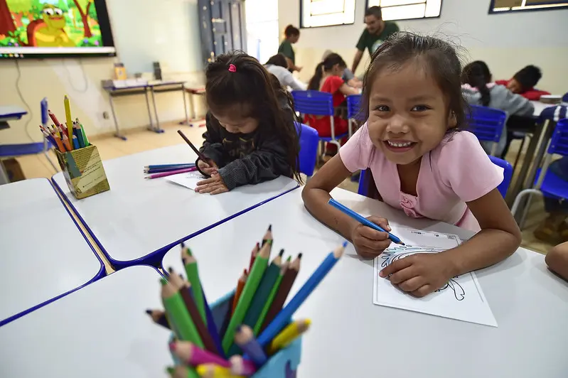 Sala de aula repleta de crianças participando de uma oficina de desenho. Em primeiro plano, uma menina sorri, enquanto segura o lápis de cor e o papel.