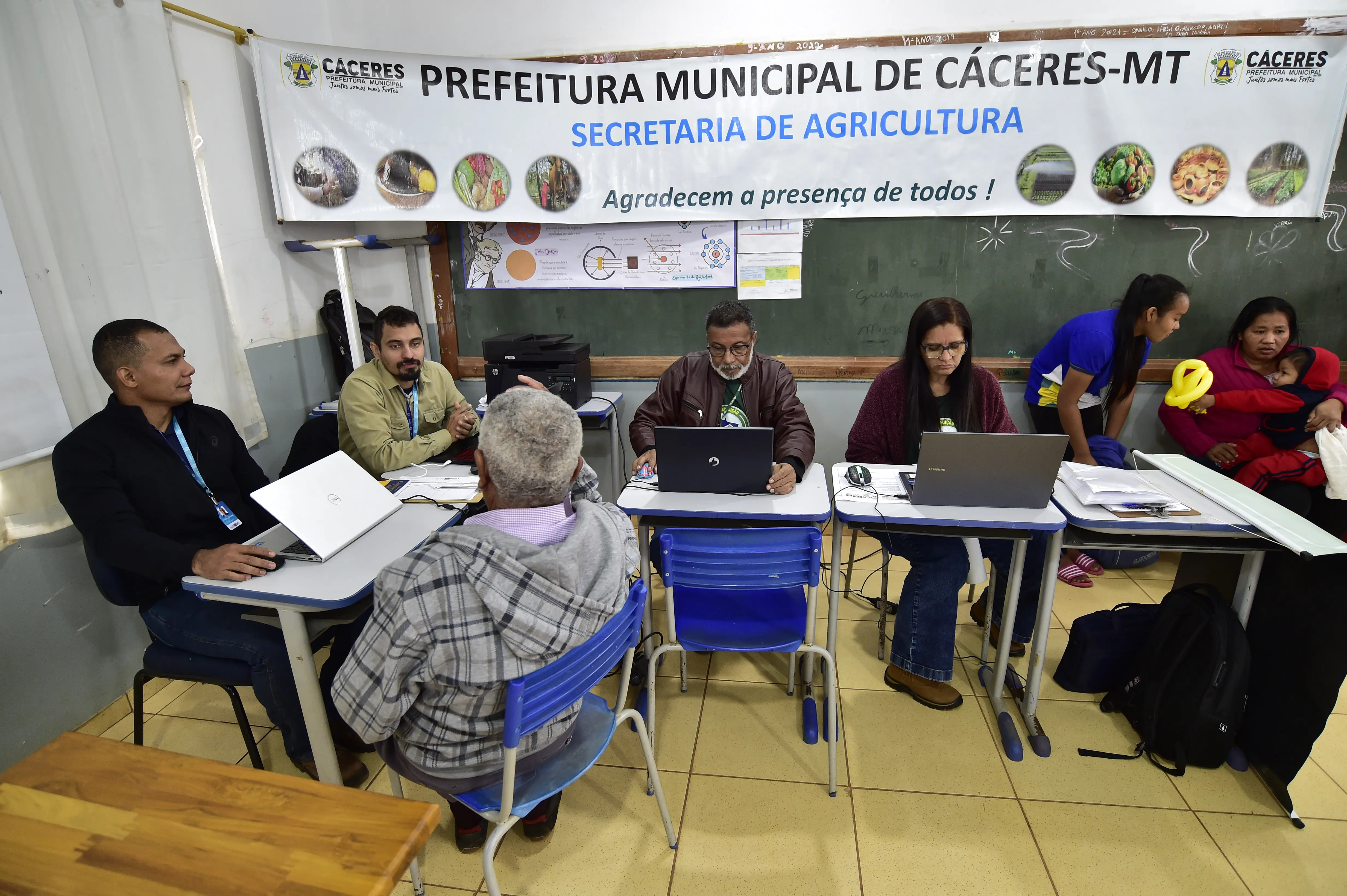 Quatro servidores atendem cidadãos com notebooks sob banner da Secretaria de Agricultura de Cáceres-MT. Mulher com bebê e jovem com balão colorido aparecem à direita da imagem.