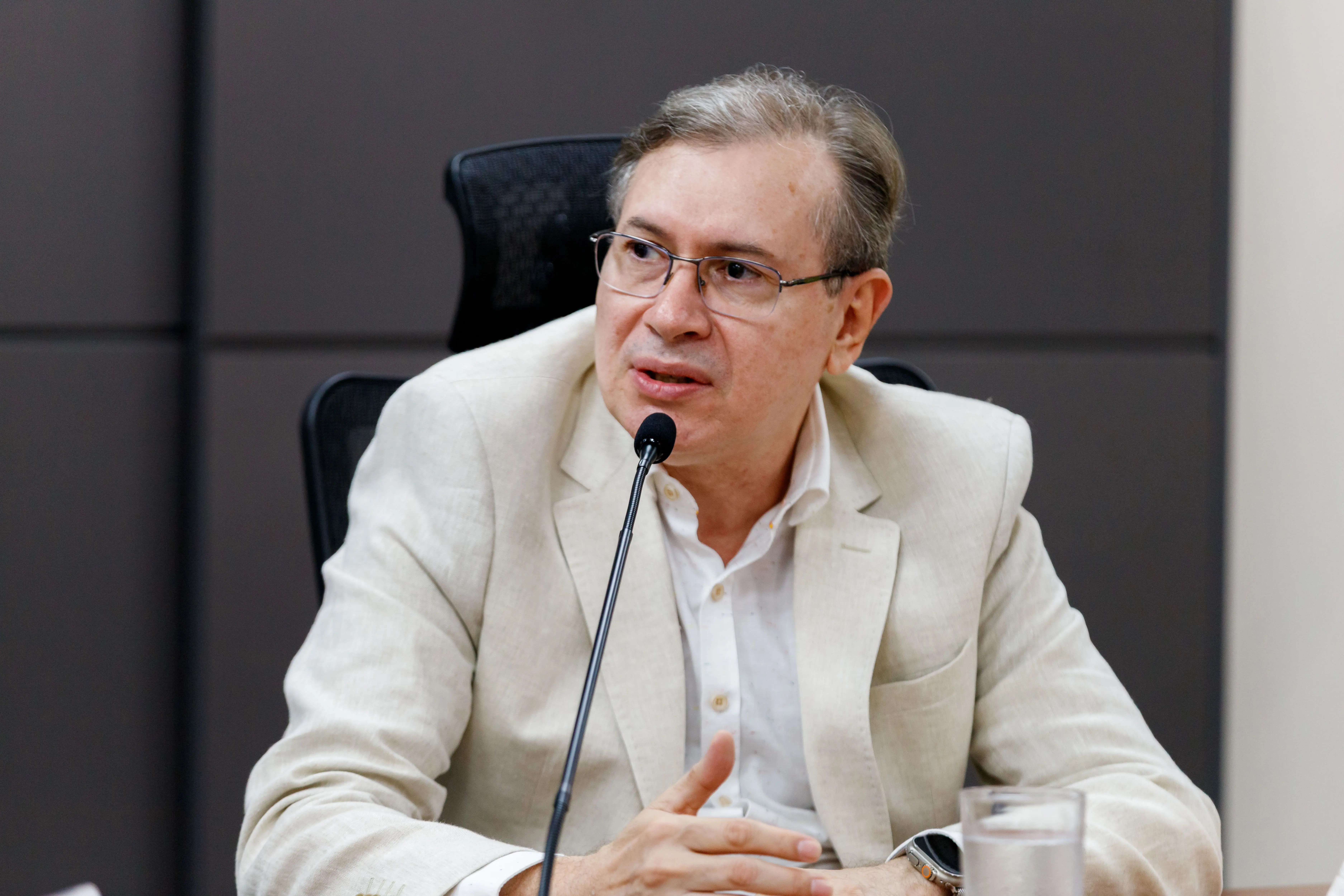O desembargador Sebastião de Arruda Almeida fala ao microfone da mesa de reuniões. Ele é um homem branco e usa óculos de grau. Veste paletó bege claro e camisa branca. O fundo é composto por painéis escuros.