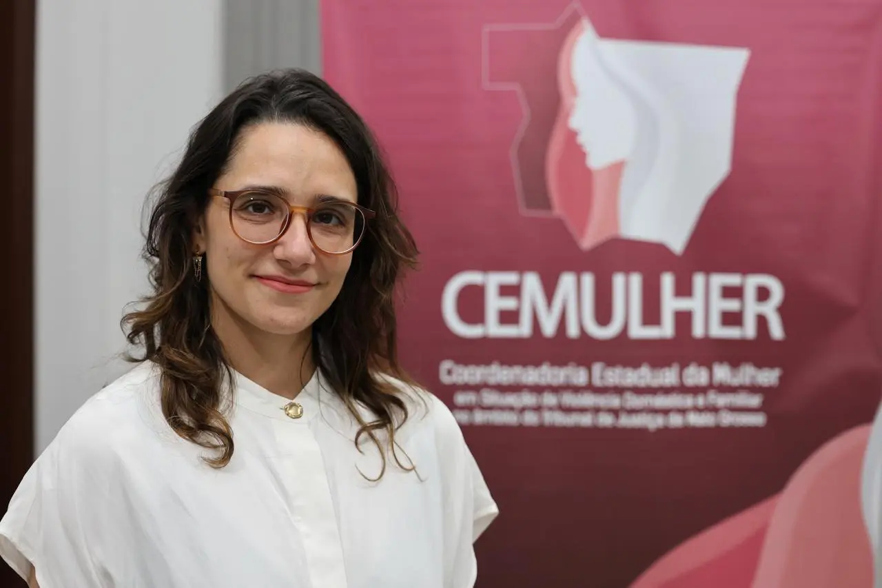 Retrato profissional de mulher de blusa branca e óculos posando em frente ao banner institucional do CEMULHER (Coordenadoria Estadual da Mulher).