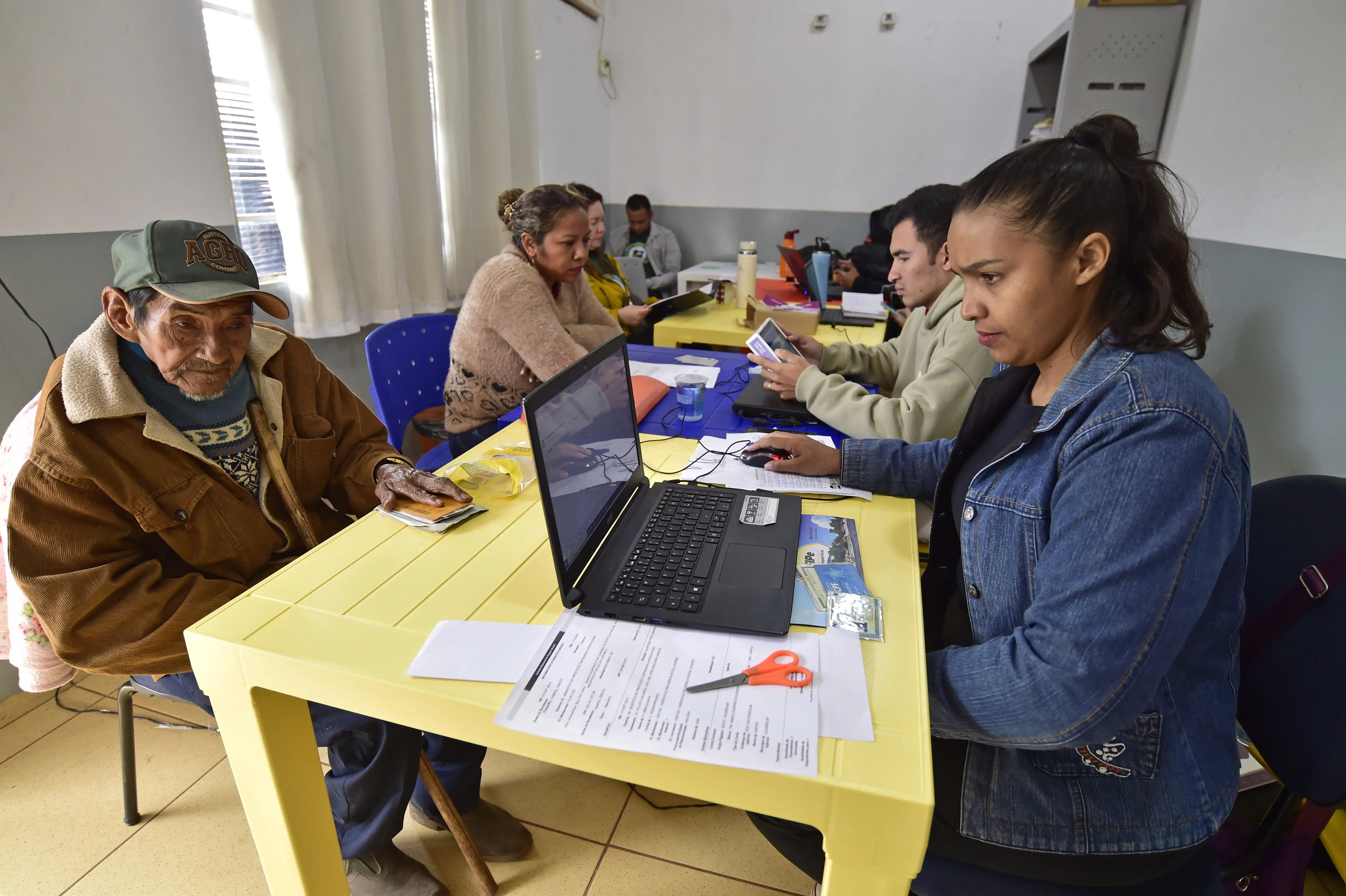 Em sala com mesas amarelas, homem idoso com boné é atendido por mulher usando notebook. Outras pessoas também são atendidas ao fundo, em ambiente iluminado e organizado.