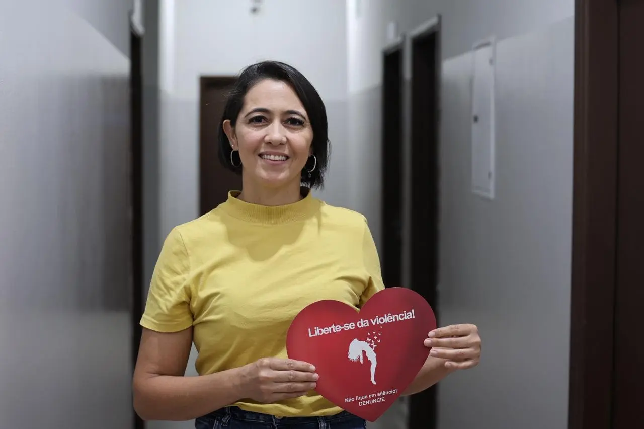 Mulher com  blusa amarela segura um cartaz vermelho em forma de coração. Ela tem cabelos curtos, pretos e sorri para foto