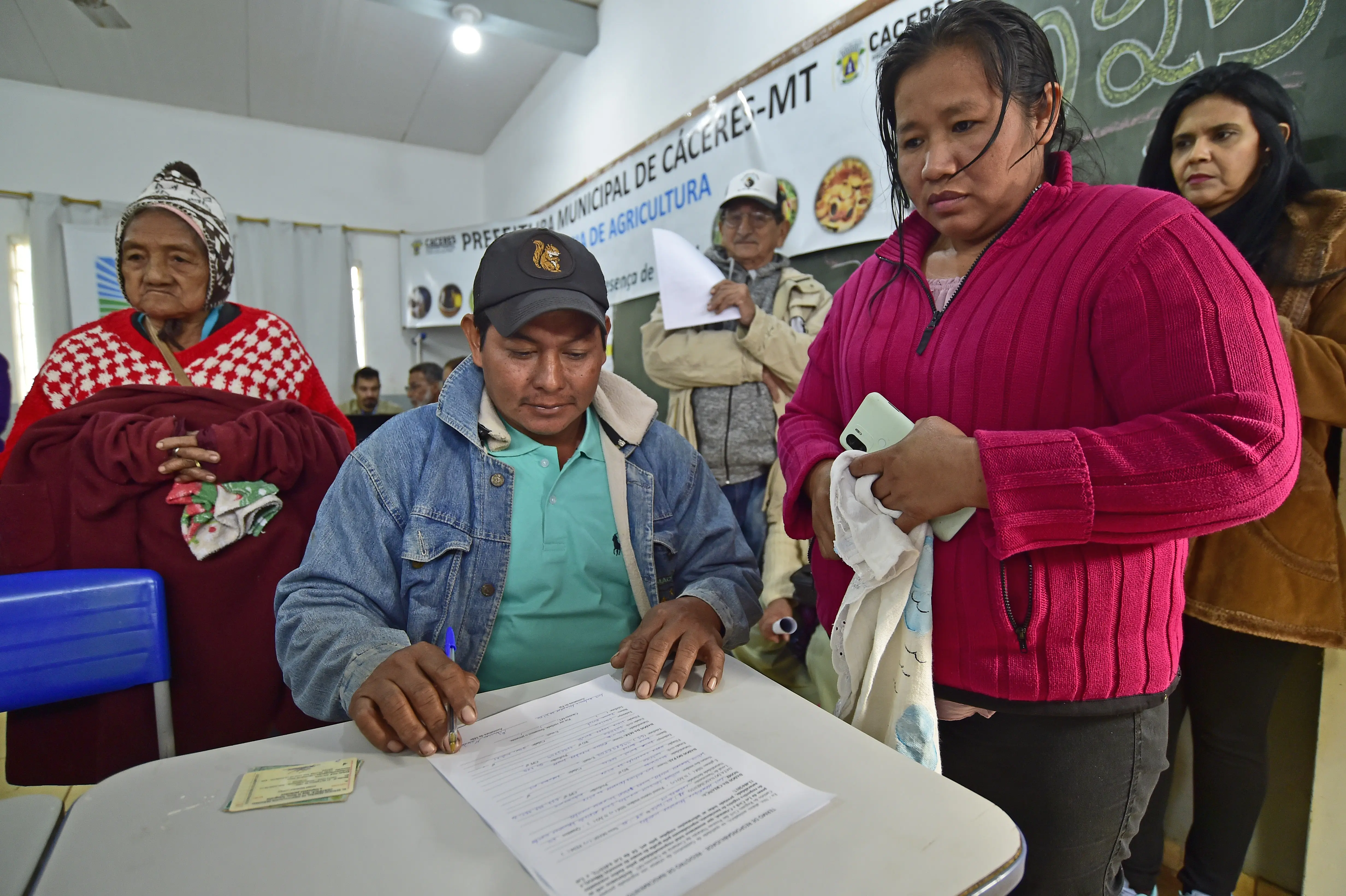 Homem de boné e camisa verde assina documento sobre mesa. Ao lado, mulher de blusa rosa segura celular e panos. Ao fundo, idosa com poncho vermelho e banner da prefeitura.