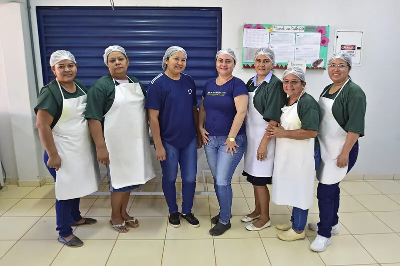 Foto que mostra sete mulheres que trabalham na cozinha da Escola 11 de Agosto posando para a foto e sorrindo.