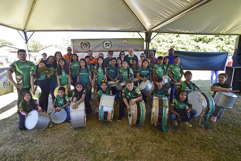 Foto que mostra os membros da fanfarra da Escola Municipal Ponta do Aterro posando e sorrindo para a foto, num gramado, embaixo de uma tenda na parte externa da escola.