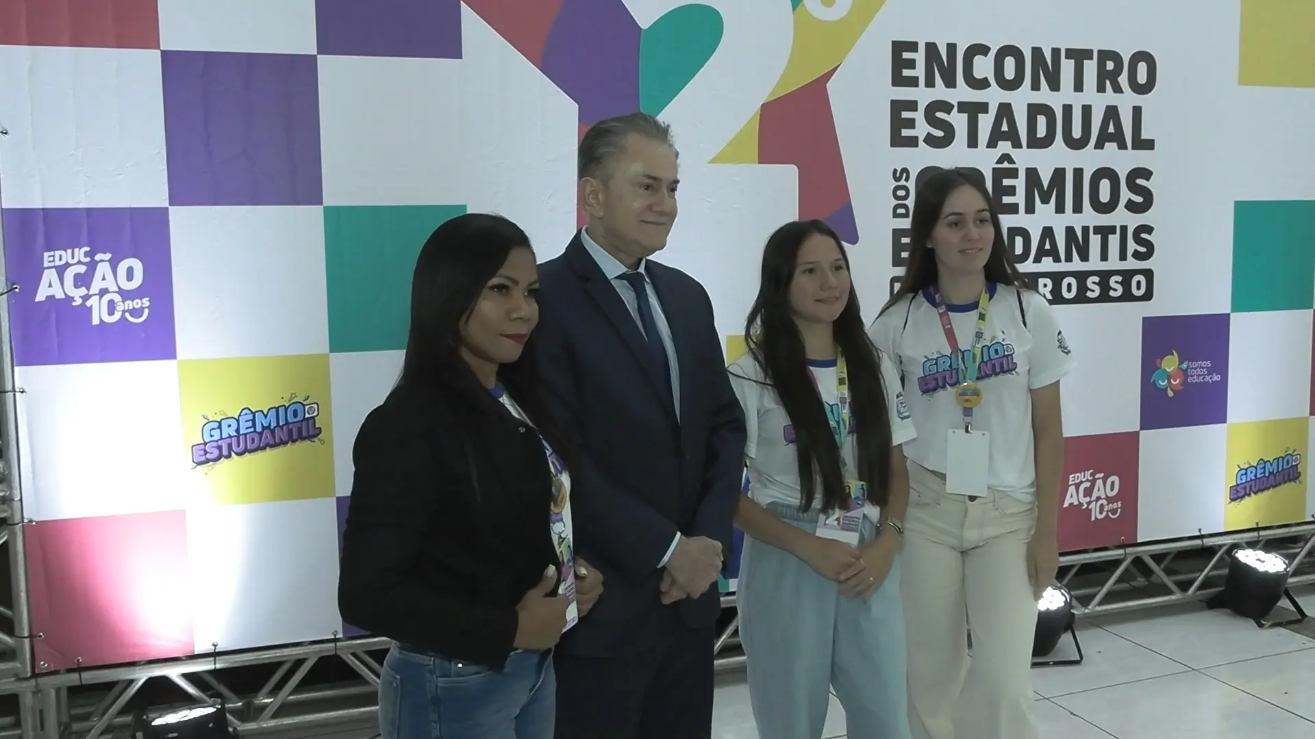 Imagem mostra autoridades e estudantes posando para foto durante o Encontro Estadual dos Grêmios Estudantis de Mato Grosso. Painel colorido ao fundo destaca logotipos do evento e do programa Educação 10 anos.