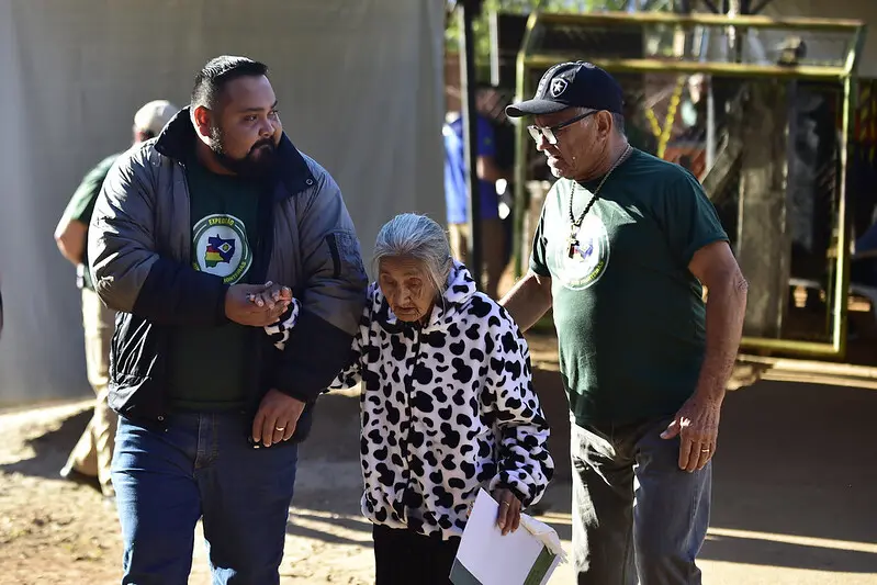Foto que mostra o coordenador da Escola Municipal Dona Lila Hill de Souza e um voluntário da Justiça Sem Fronteiras ajudando uma idosa a andar no pátio da escola.