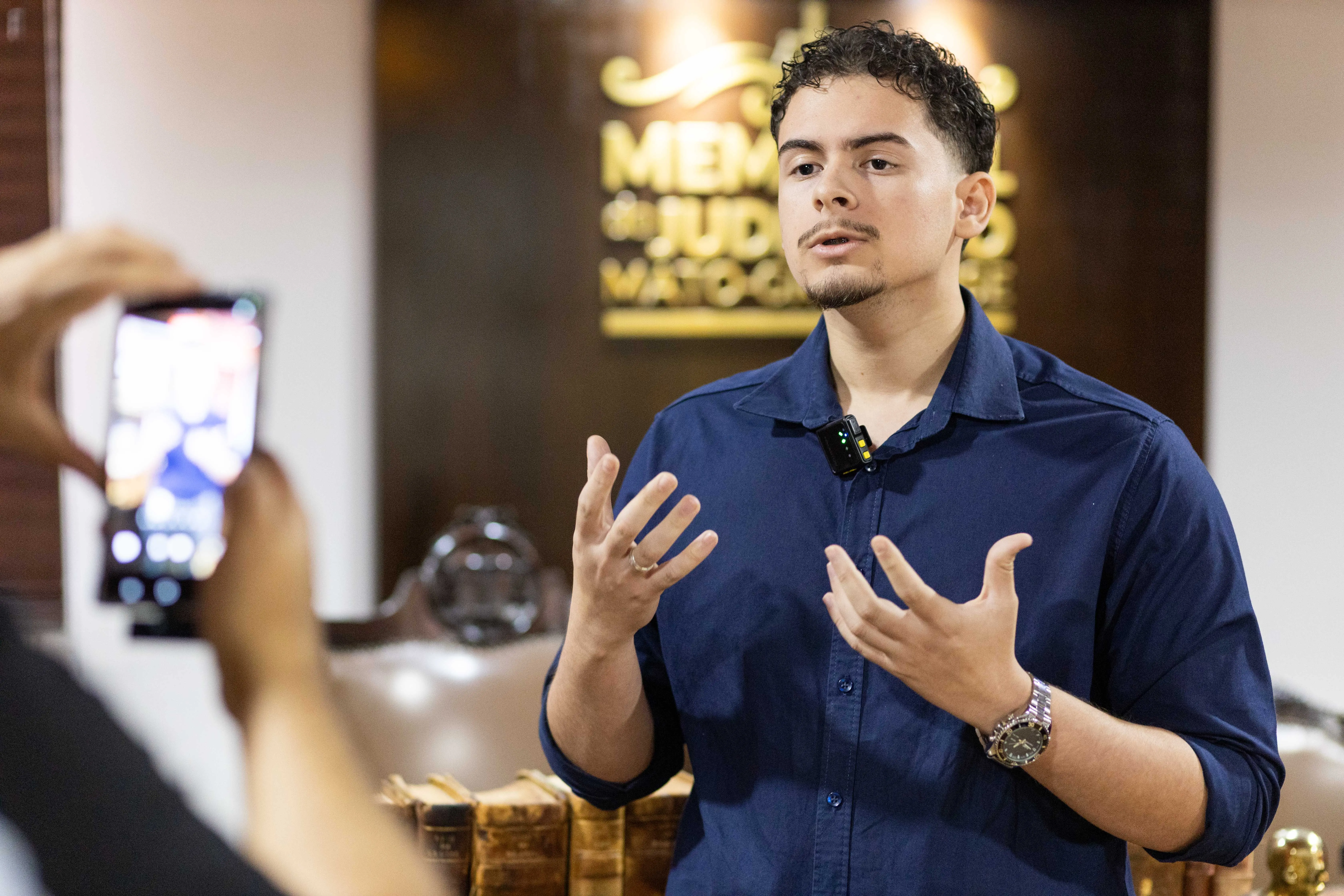 O estudante de Direito Wagner Galvão de Vasconcelos, um jovem de cabelos e olhos escuros e barba curta, veste uma camisa polo azul escura e gesticula com as mãos enquanto fala. Ele usa um relógio prateado no pulso esquerdo. Ao fundo, vê-se parte de uma parede de madeira com a inscrição "Memorial do 