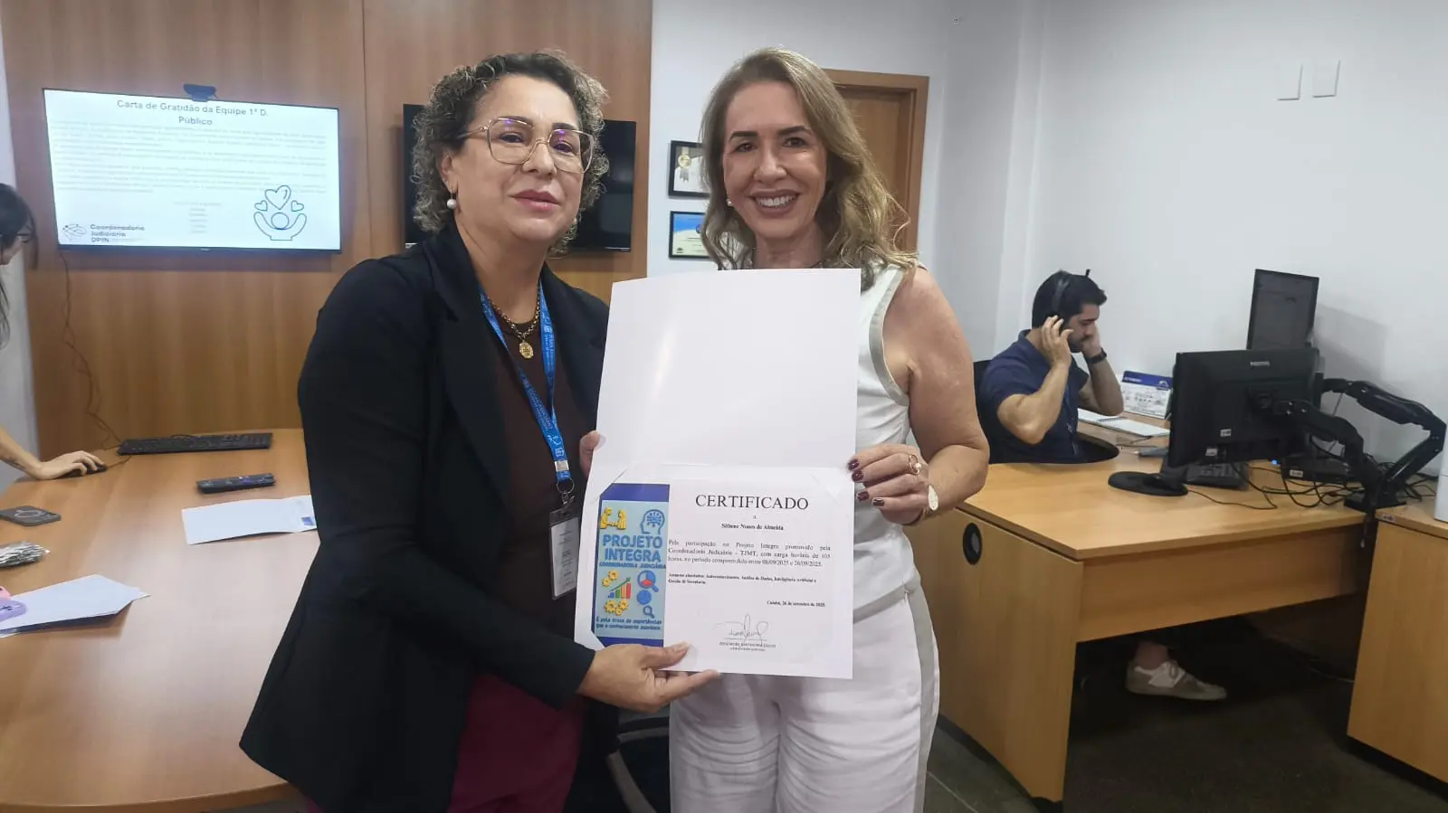 Onze pessoas posando com certificados, vestindo tons pastéis. Vários usam crachás. Ao fundo, parede branca com porta de madeira e TV com conteúdo azulado.